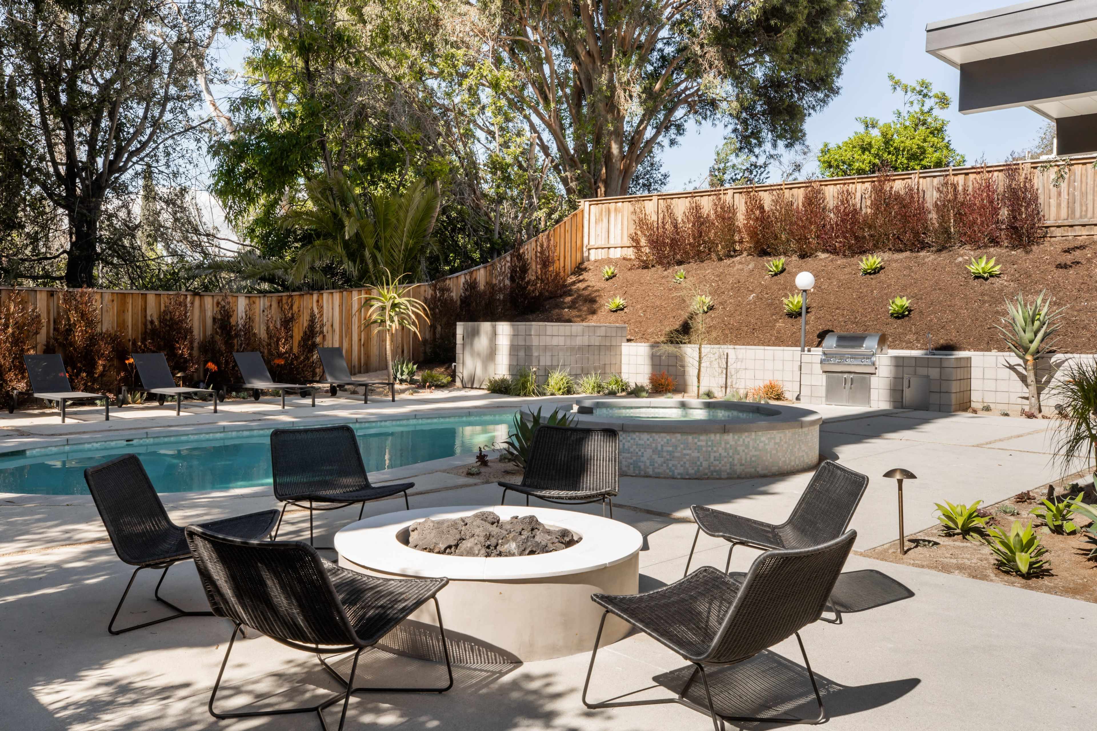 A modern outdoor space features a swimming pool, spa, and a circular fire pit surrounded by black lounge chairs.