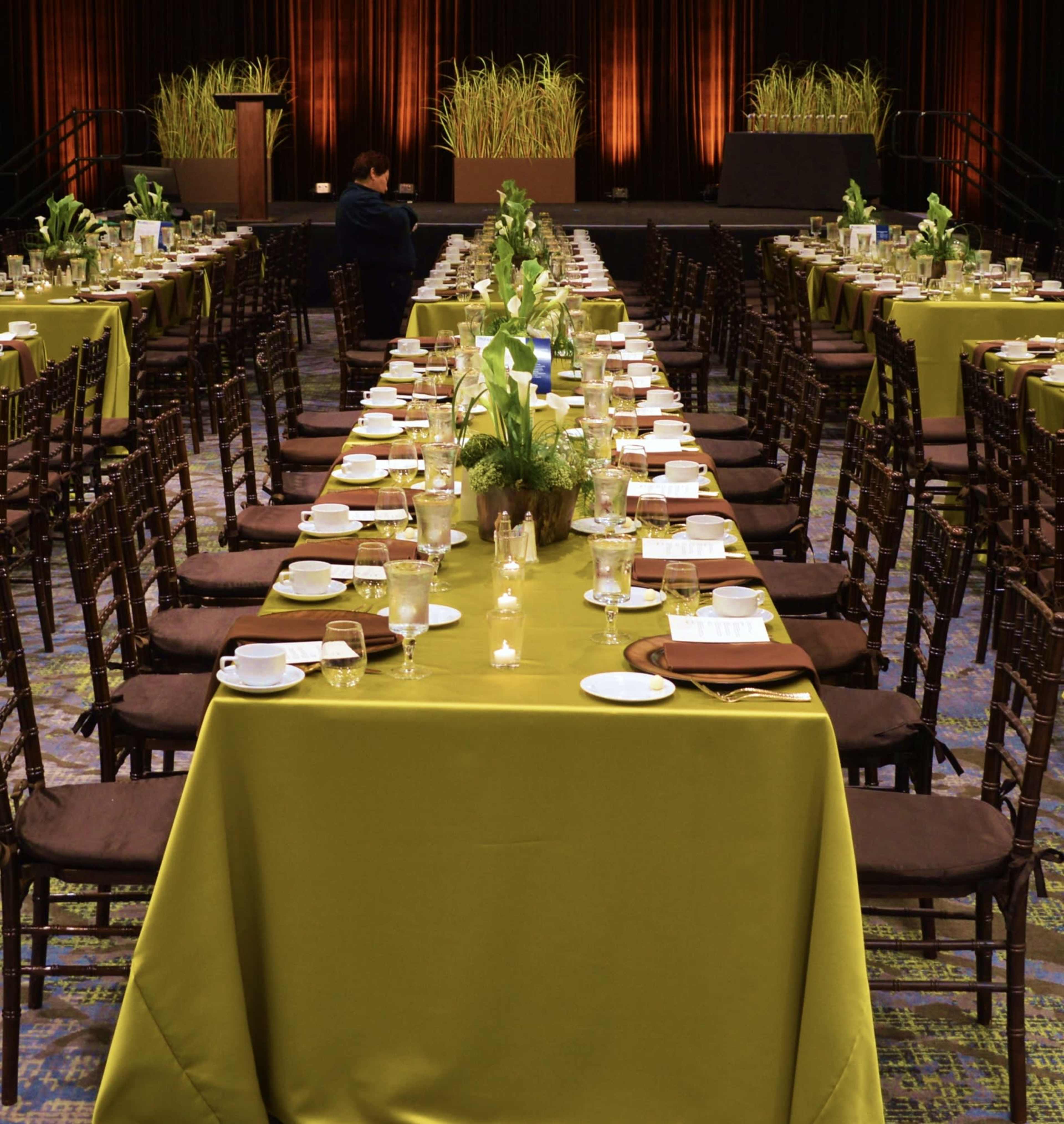 A large banquet hall is set with multiple long tables covered in green tablecloths, arranged with plates, cups, and centerpieces of plants.