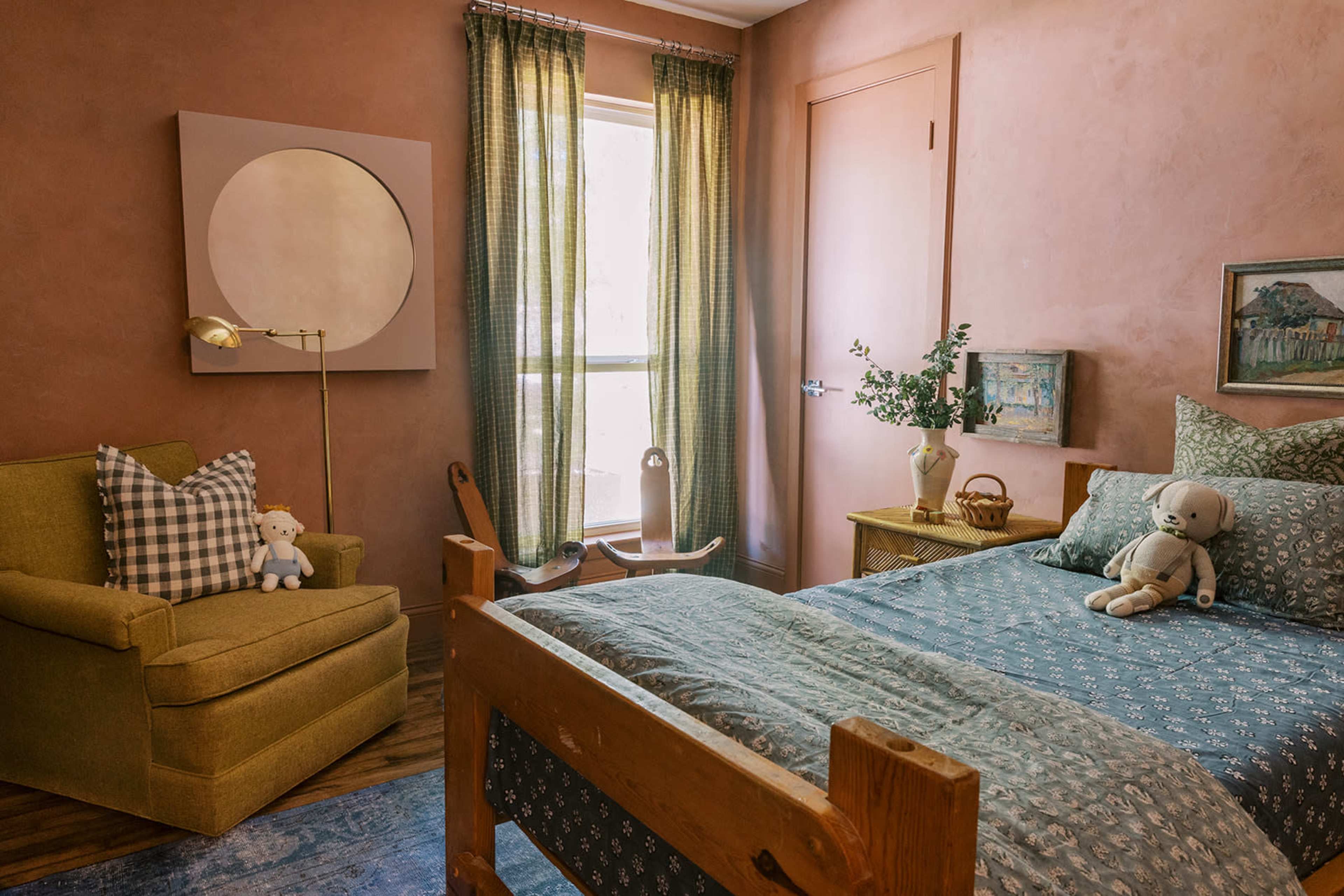 A cozy bedroom features a wooden bed with patterned bedding, a green armchair with a stuffed toy, and a window dressed with curtains, allowing natural light to fill the space.