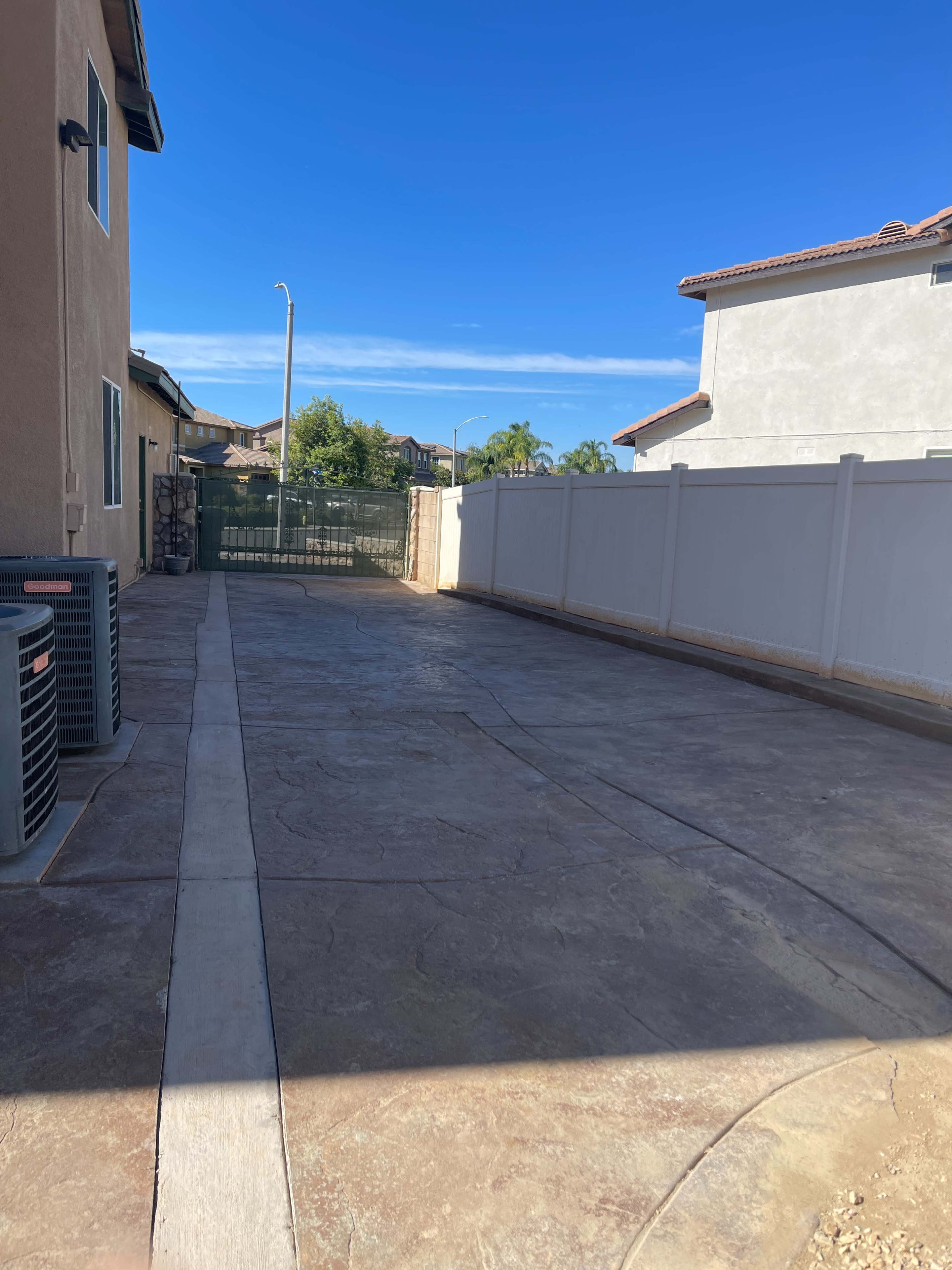The image shows a wide, paved driveway between two residential buildings, with a tall white fence and a green gate in the background.