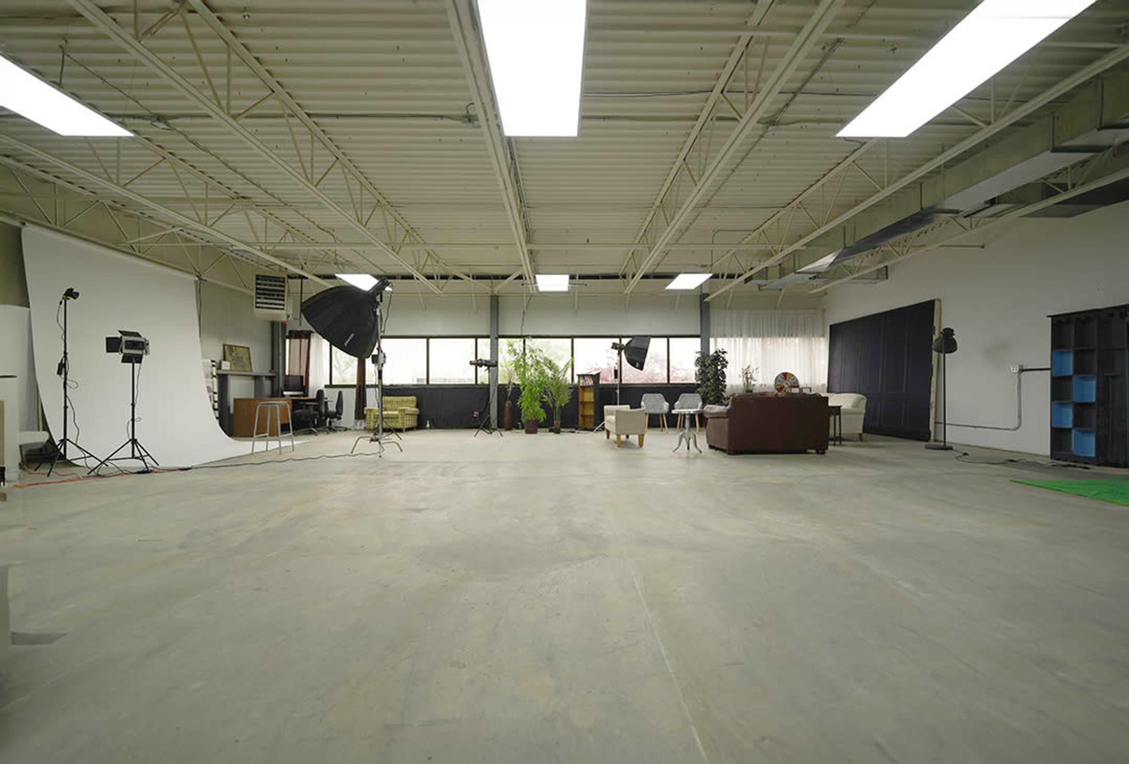 A spacious studio with a concrete floor, large windows, and various lighting equipment set up alongside furniture and backdrops.