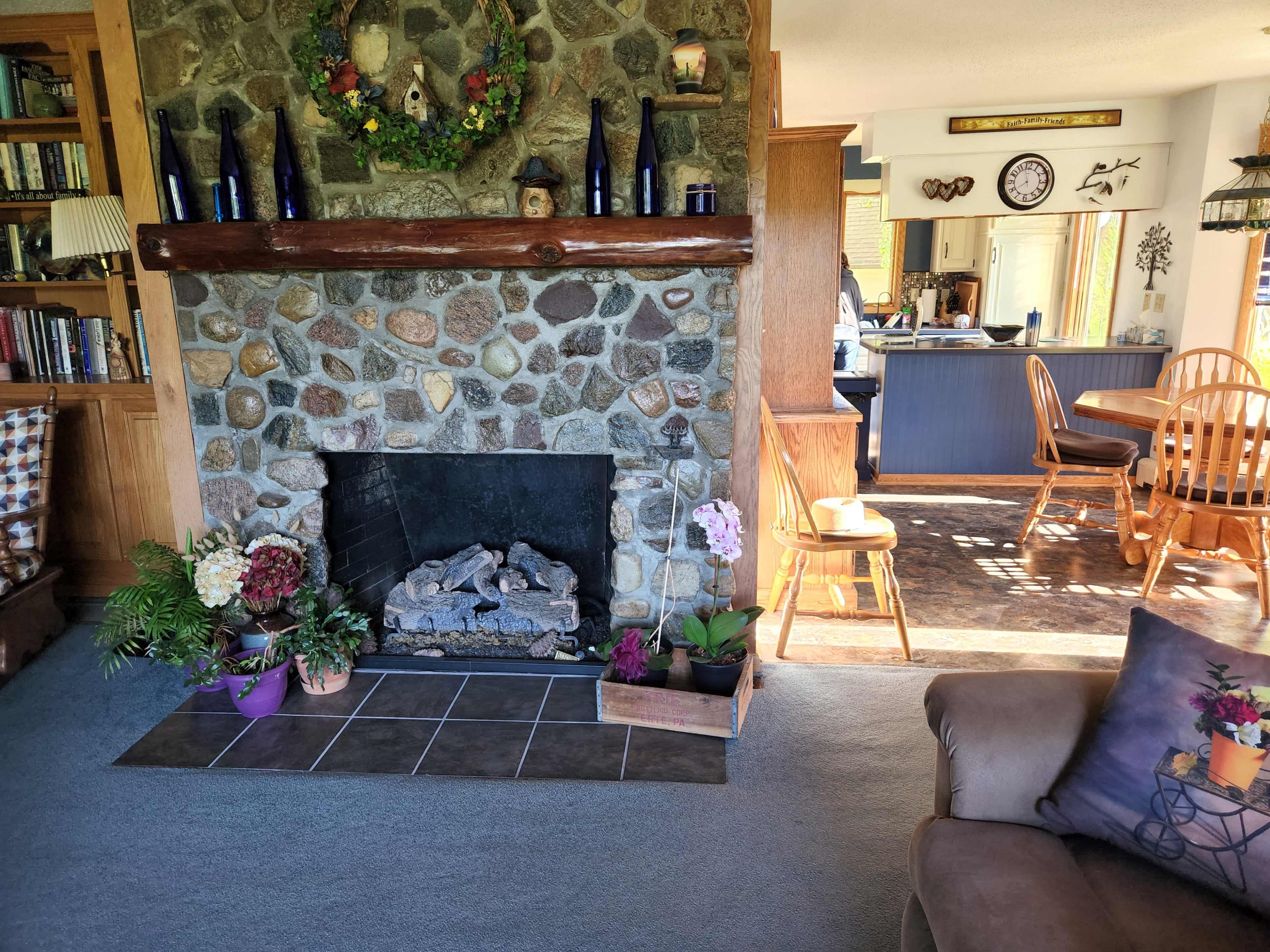 A living room with a stone fireplace, surrounded by plants and decorative items, leading to a kitchen and dining area in the background.