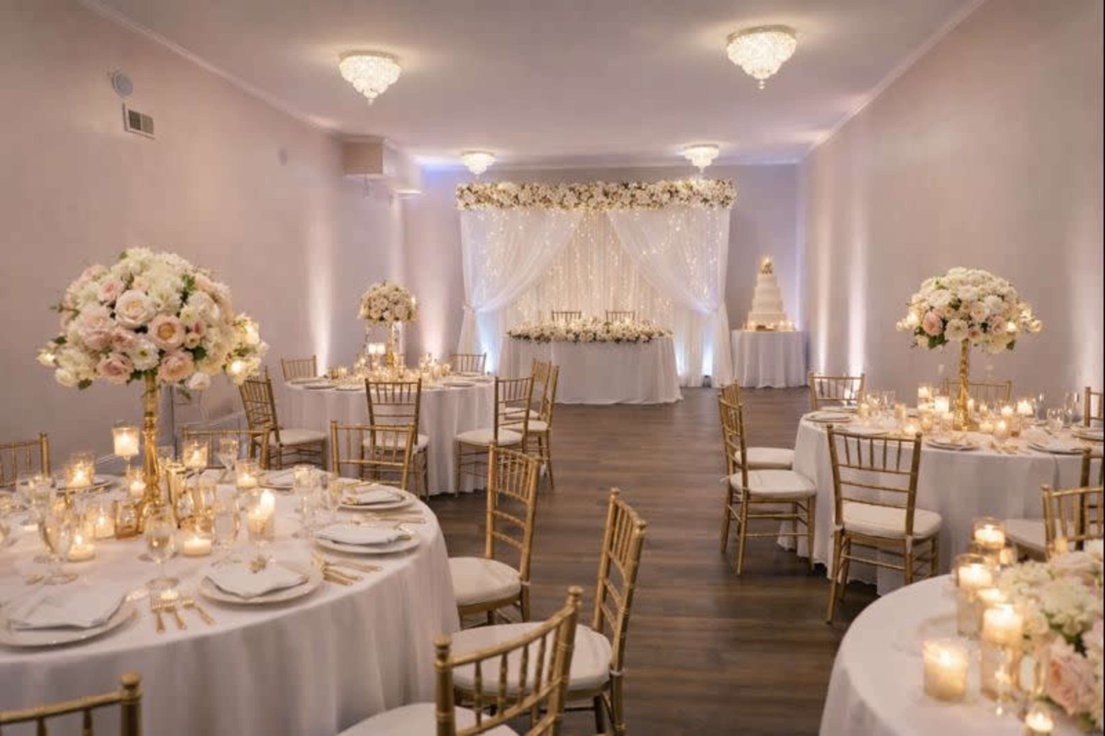 The image shows a beautifully arranged wedding reception with round tables adorned with floral centerpieces, soft lighting, and a decorated backdrop featuring a cake and candles.