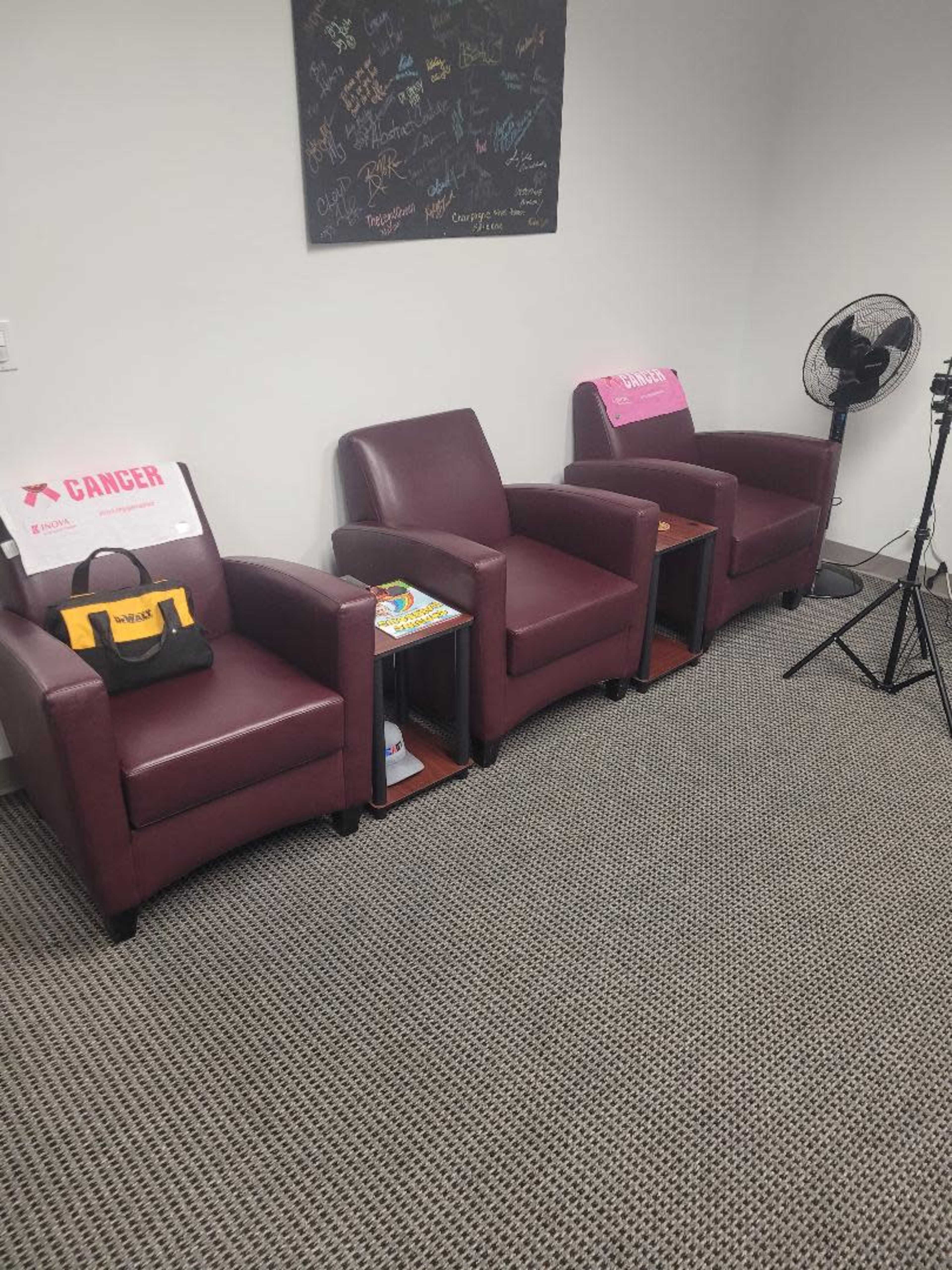 The image shows three maroon chairs arranged against a wall with a fan and a small side table between them, accompanied by a stack of magazines and a bag on one of the chairs.