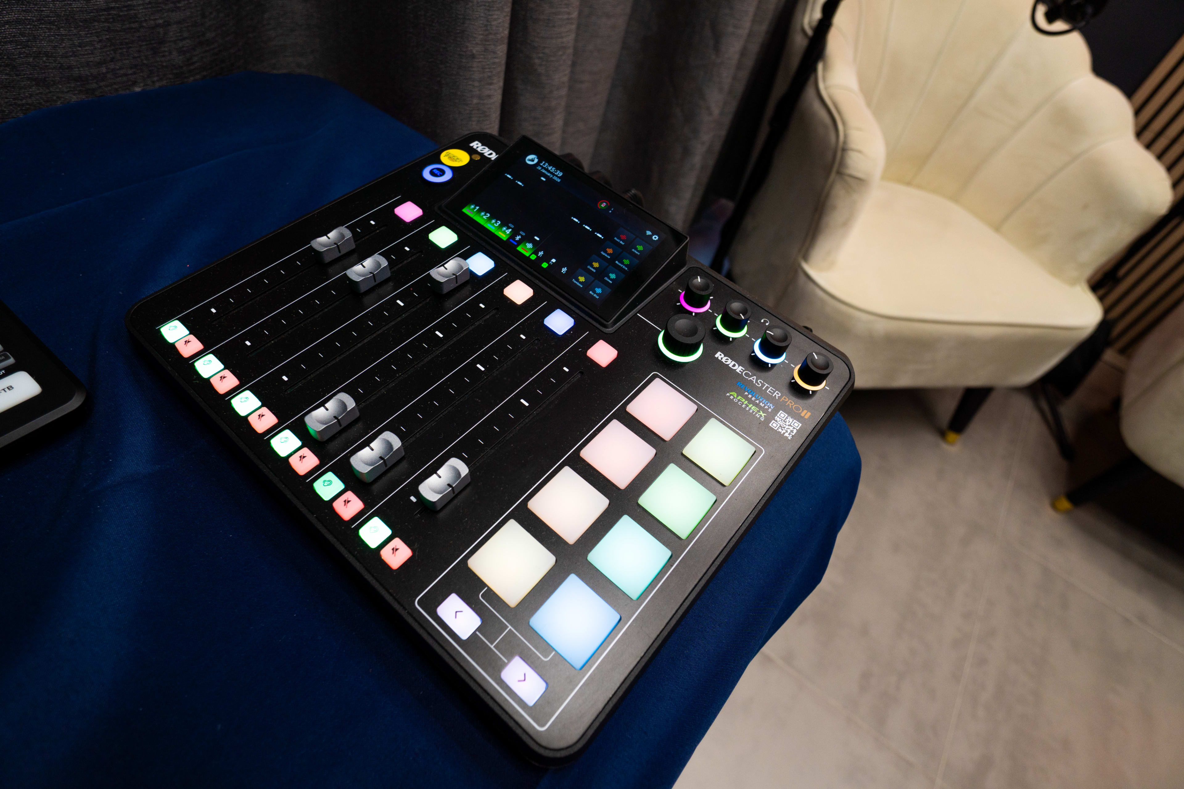 A digital audio mixer with various faders, buttons, and a screen is placed on a blue table next to a cream-colored chair.