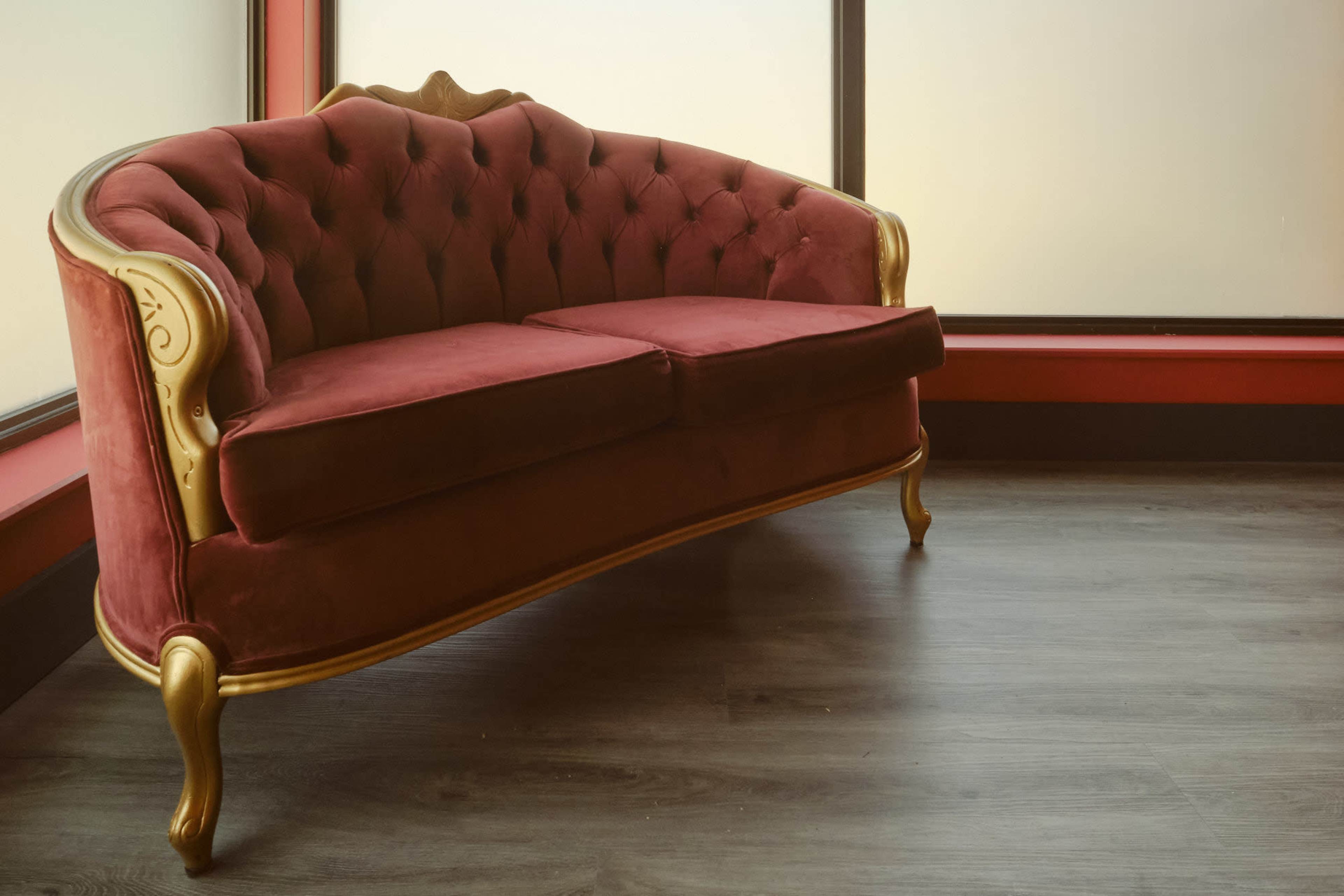 A red velvet tufted sofa with gold accents is positioned near a large window in a room with a wooden floor.