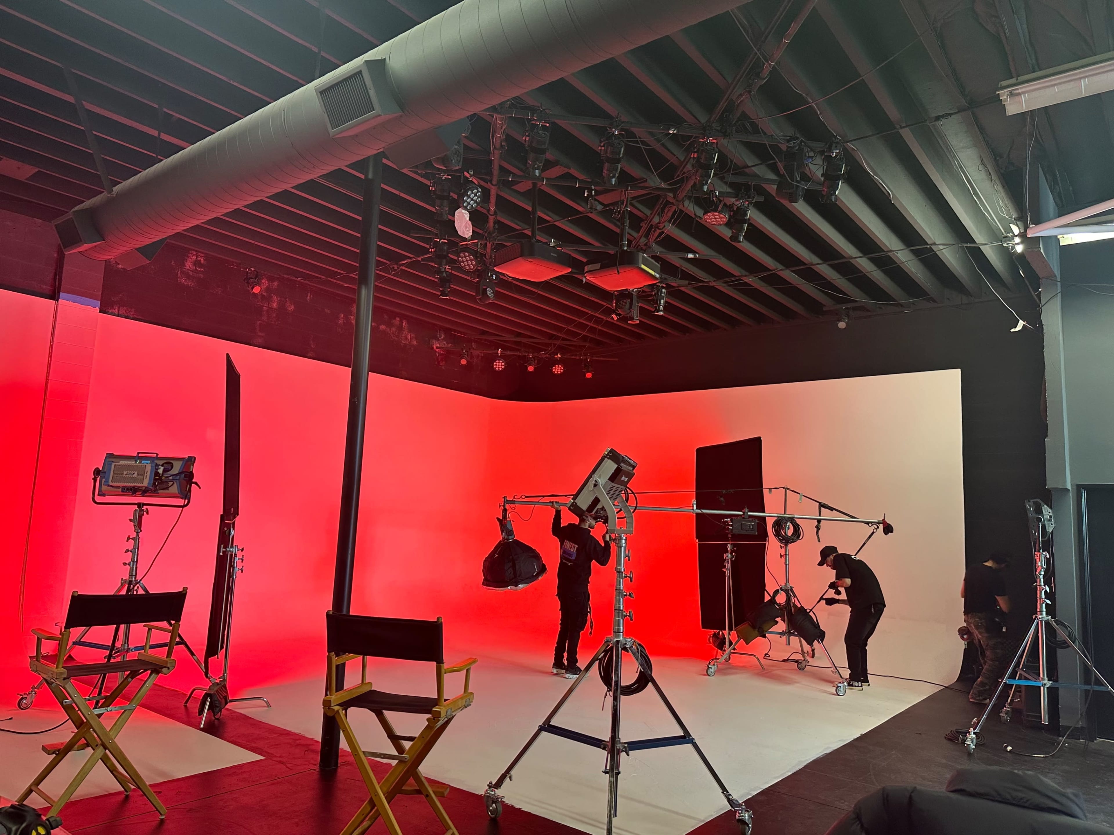 A film studio is set up with multiple lighting rigs and crew members preparing for a shoot against a red and white backdrop.