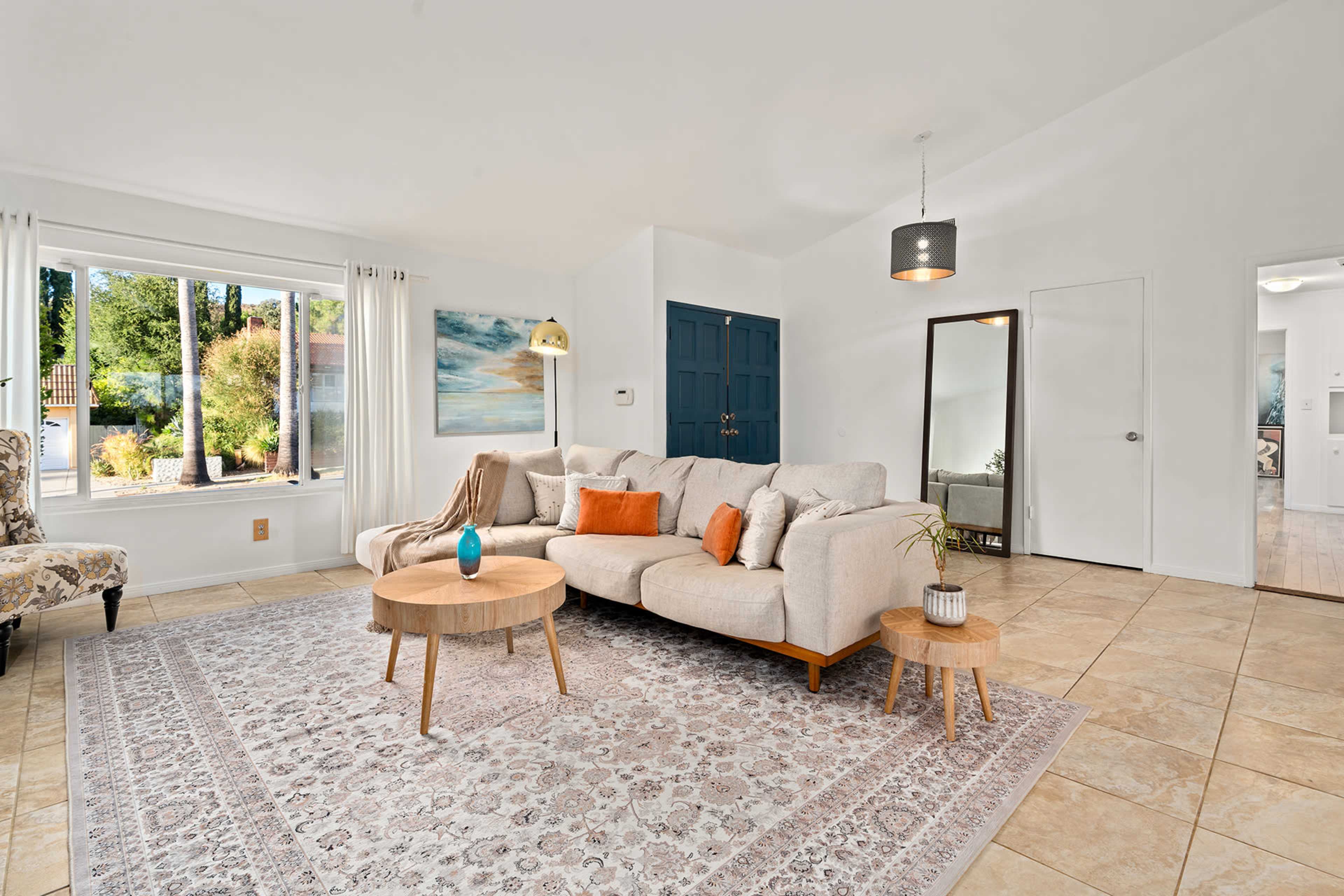 The living room features a large beige sectional sofa, a round wooden table, and a decorative rug, with large windows allowing natural light to illuminate the space.