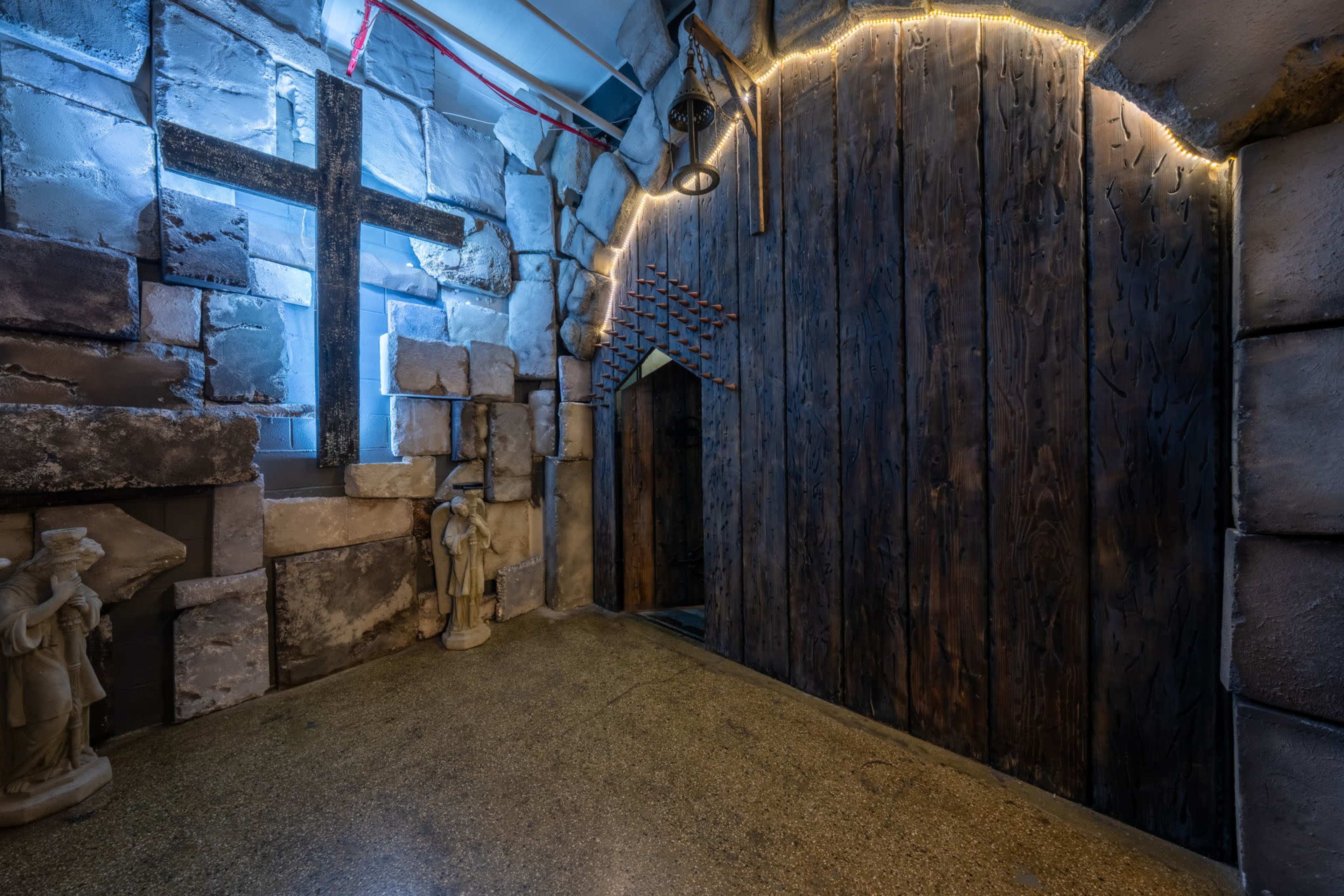 The image shows a dimly lit, stone-walled corridor featuring a large wooden door adorned with metal accents and a prominent cross illuminated by blue light, alongside two statues.