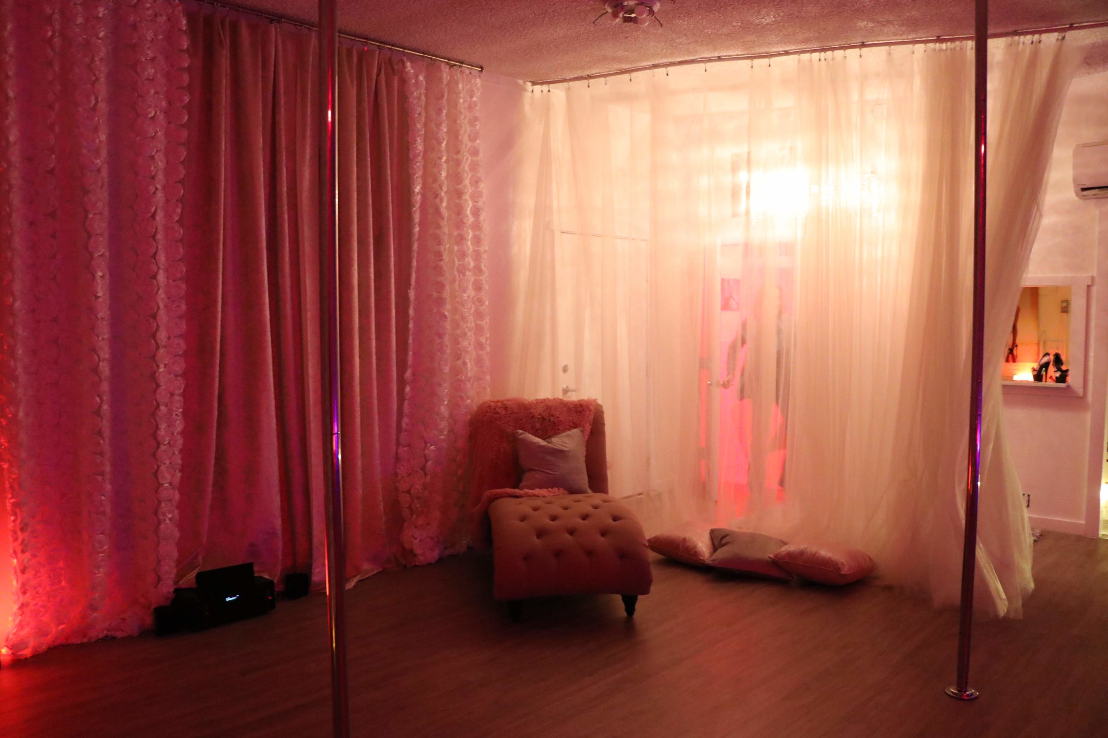 A softly lit interior room with sheer curtains, a plush chaise lounge, and two poles, suggesting a dance or performance space.