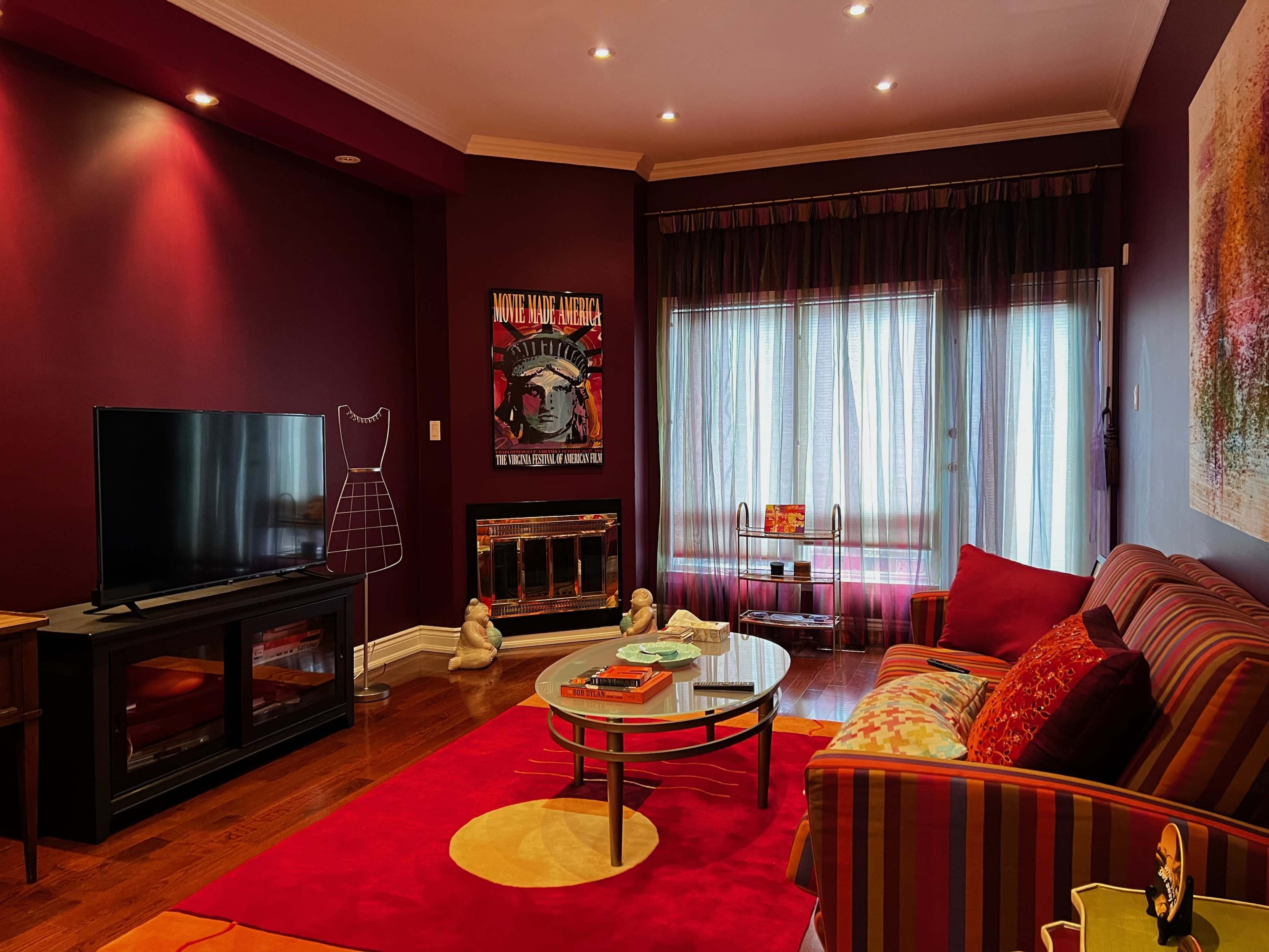 A cozy living room featuring a striped sofa, a round glass coffee table, a television, and a large window covered with sheer curtains.