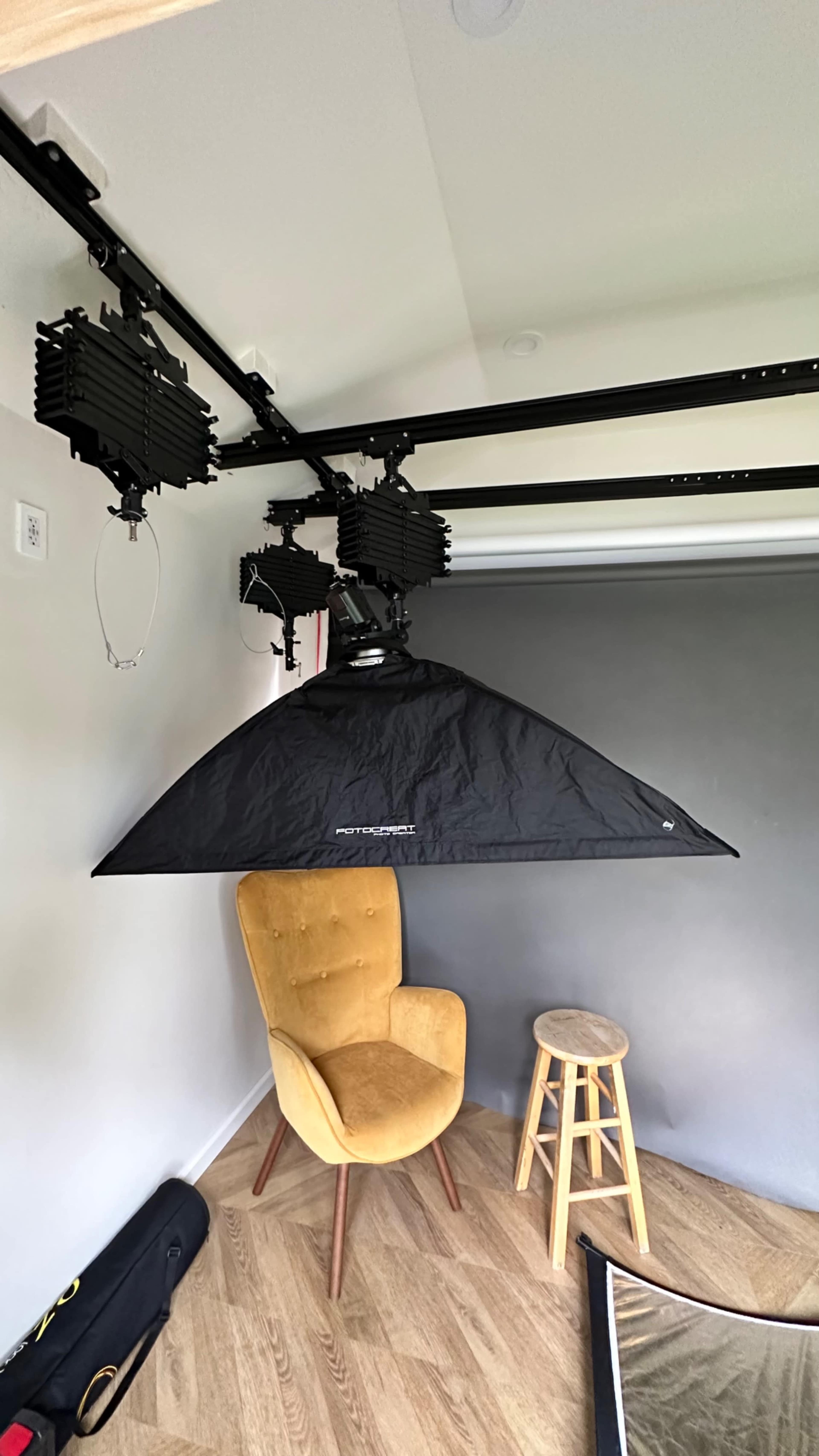 A room with a yellow armchair and a wooden stool beneath a black overhead lighting setup mounted on a track.