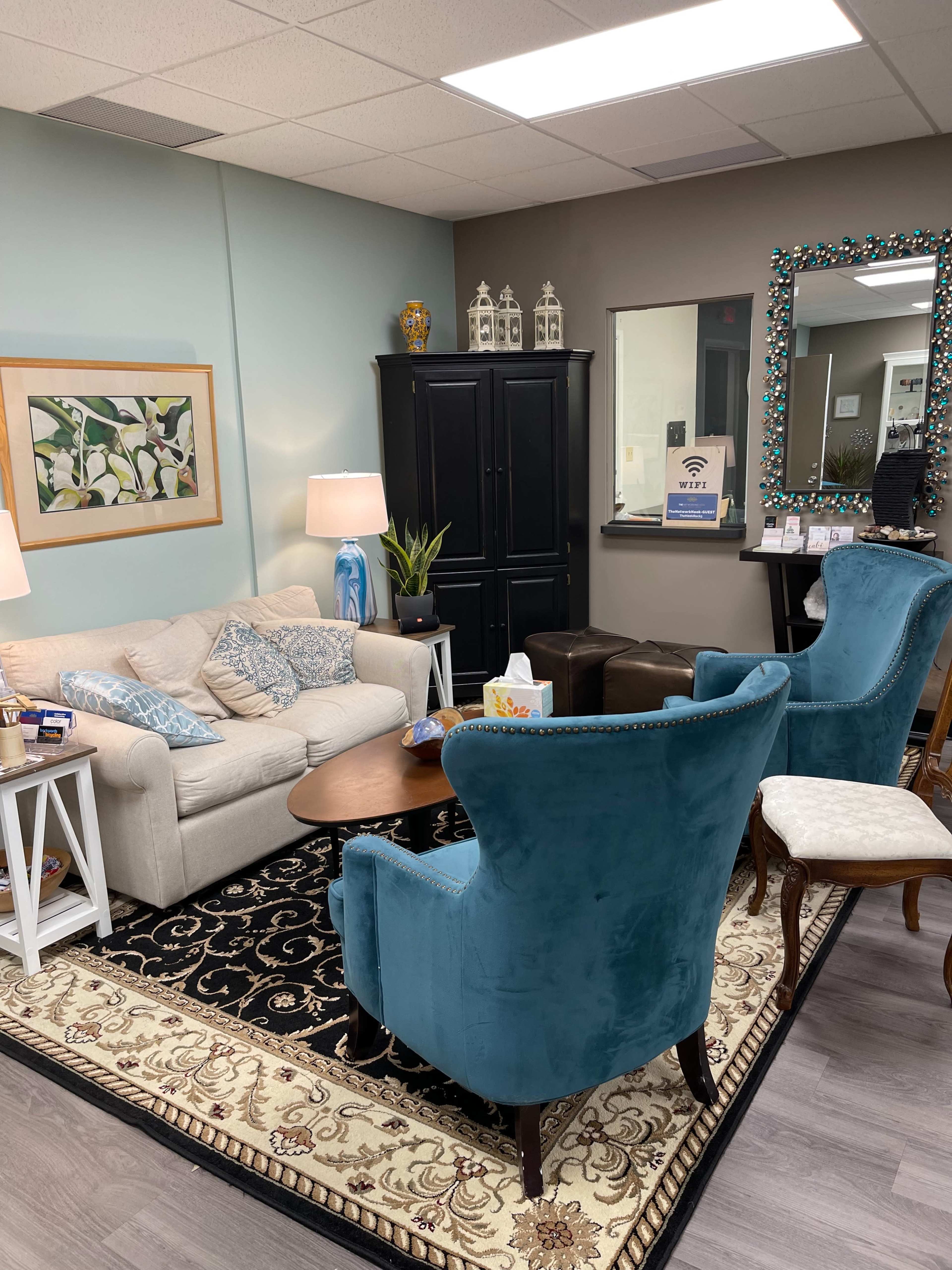A cozy waiting area with a cream-colored sofa, two blue chairs, a wooden coffee table, and decorative items on the walls and furniture.
