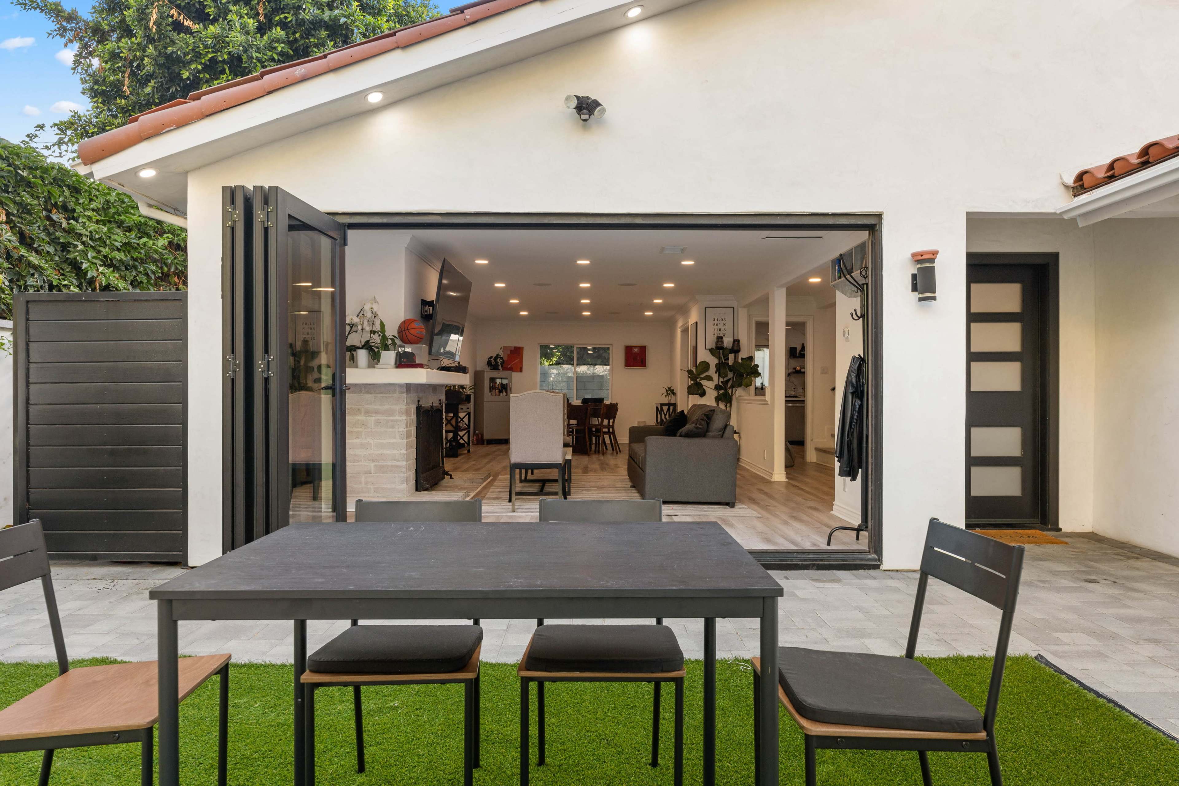 A modern outdoor space features a black table with chairs on green artificial grass, leading to an open living area with large windows and a view of the interior furnishings.