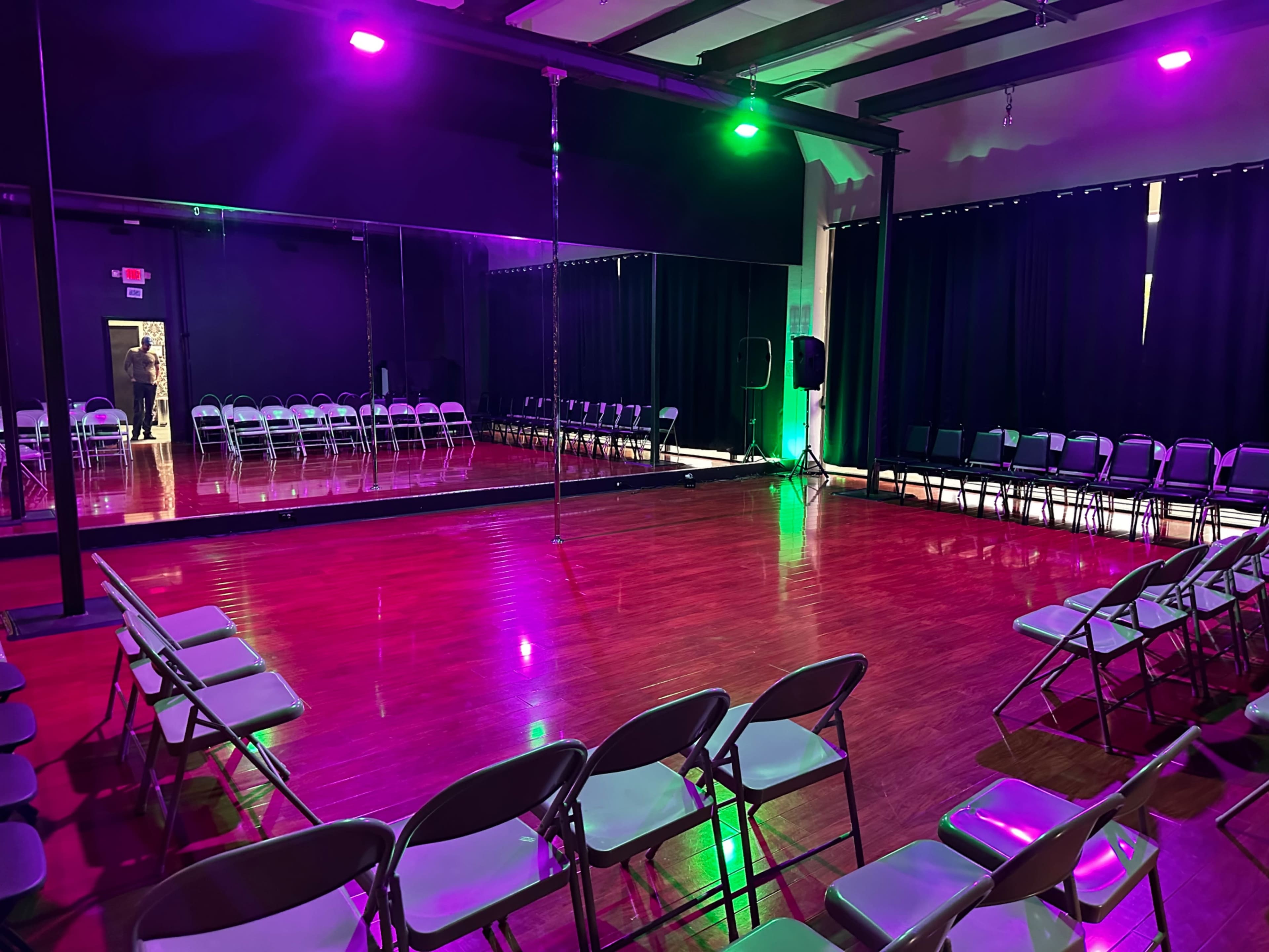 A dance studio with a polished wooden floor, a large mirror, and rows of folding chairs set up for an audience, illuminated by green and purple lights.