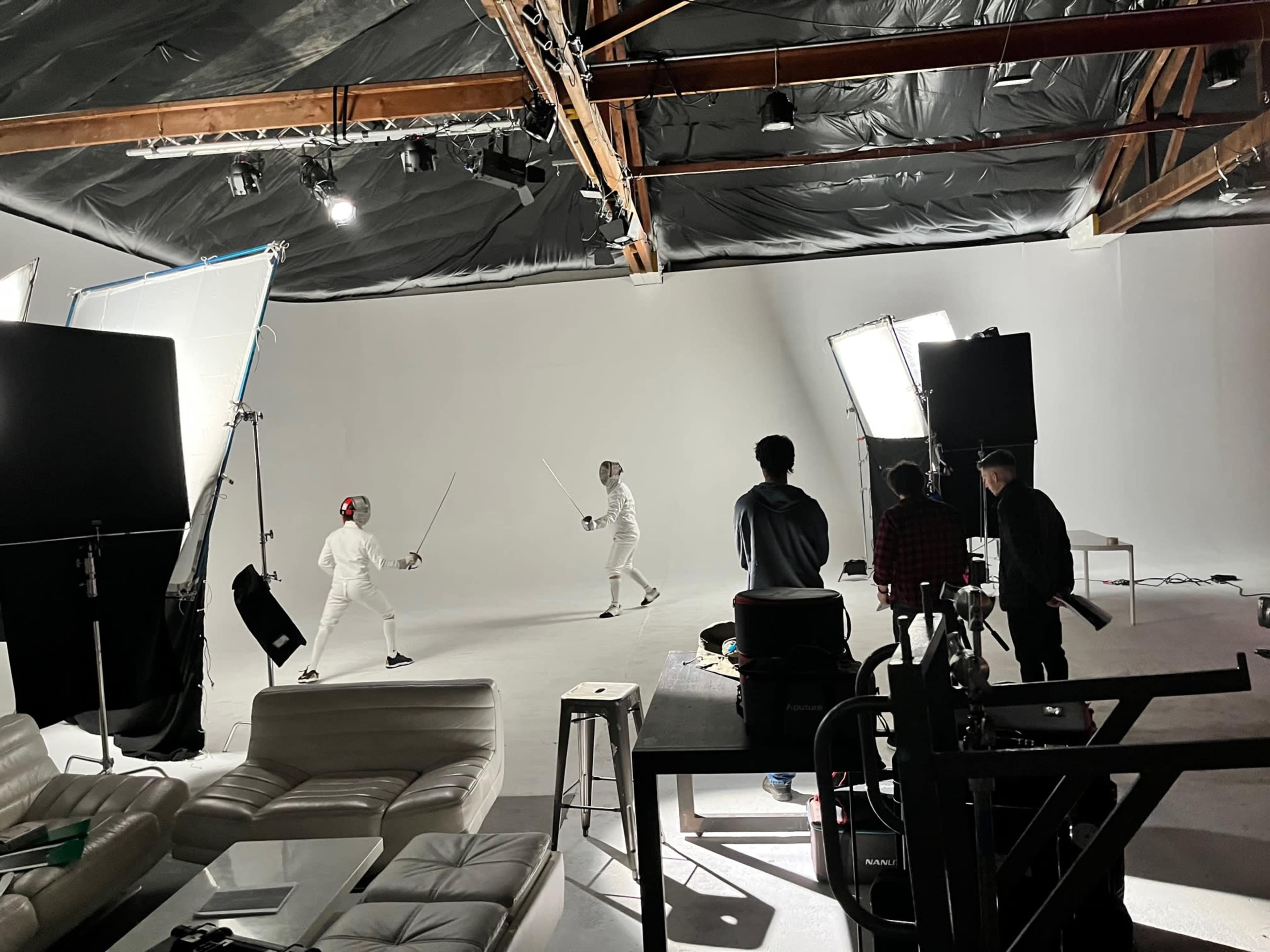 Two fencers in white gear spar in a studio setting, while crew members observe and manage lighting equipment.