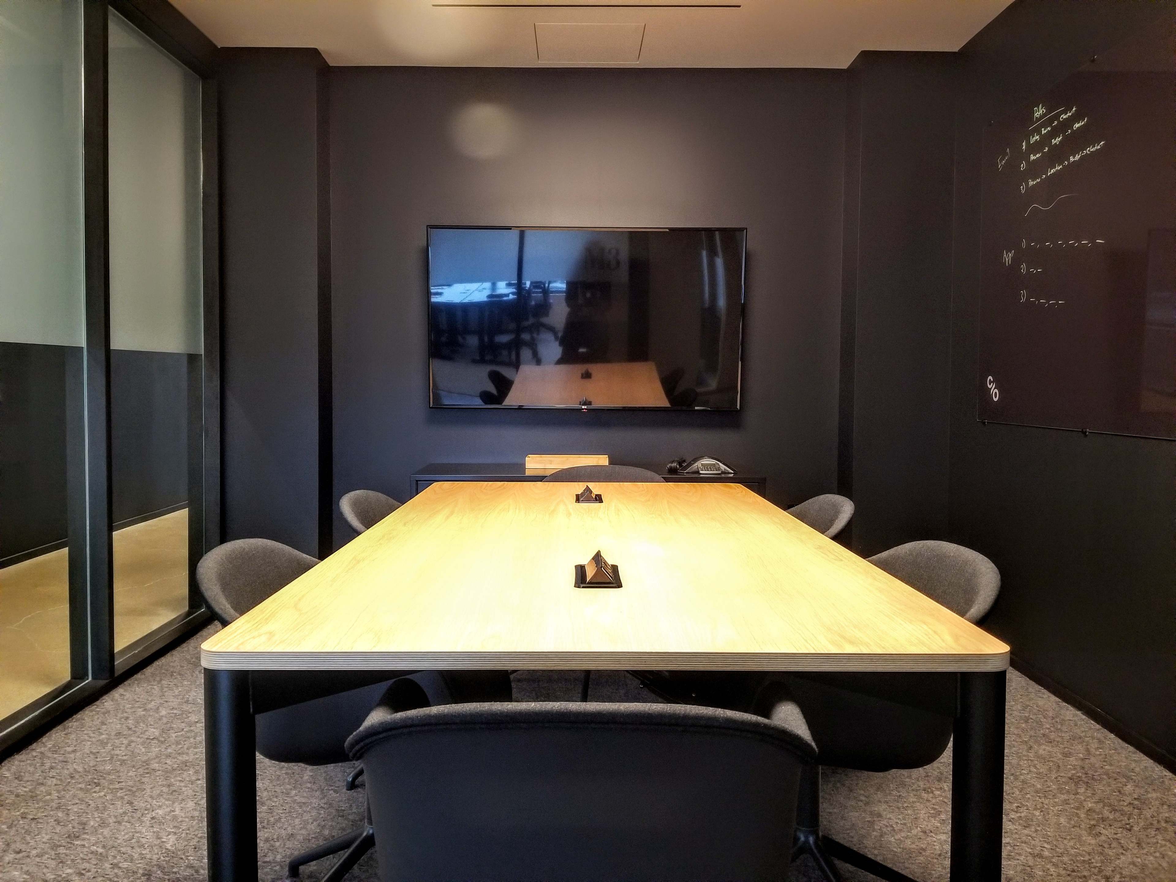 A modern conference room features a large wooden table surrounded by chairs and a flat-screen TV mounted on the wall.