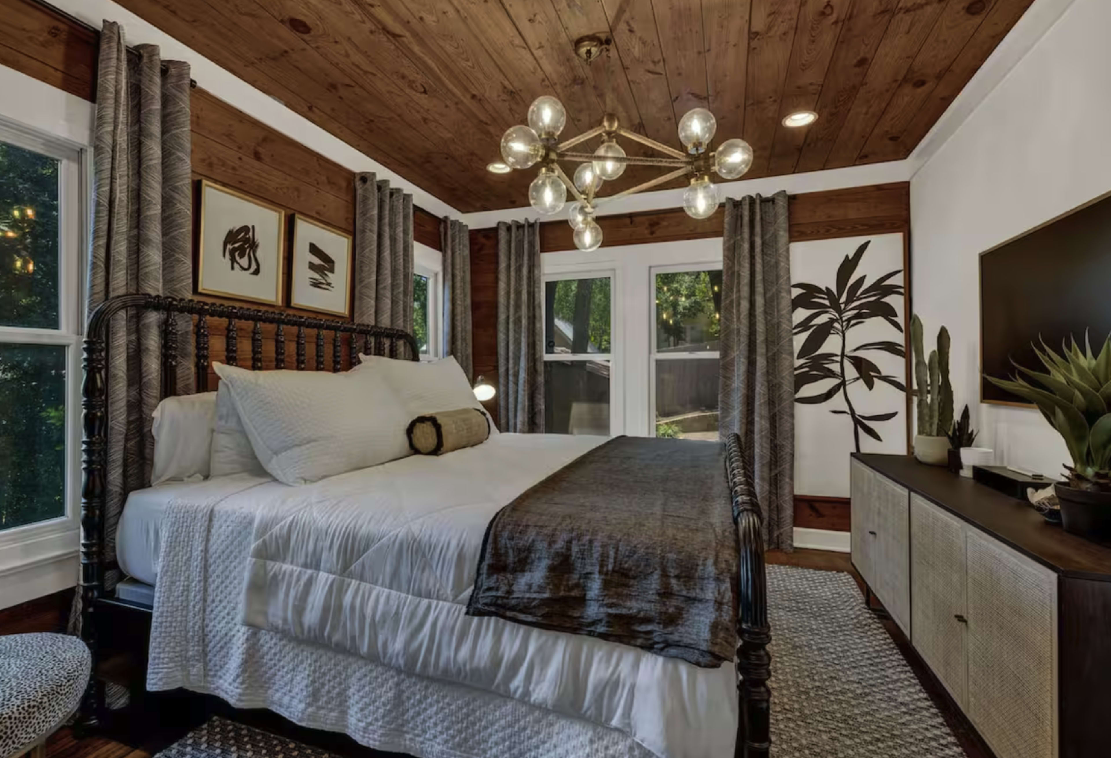 The image shows a cozy bedroom featuring a bed with white linens, a decorative chandelier, and large windows adorned with curtains, alongside a modern television cabinet and plant decor.