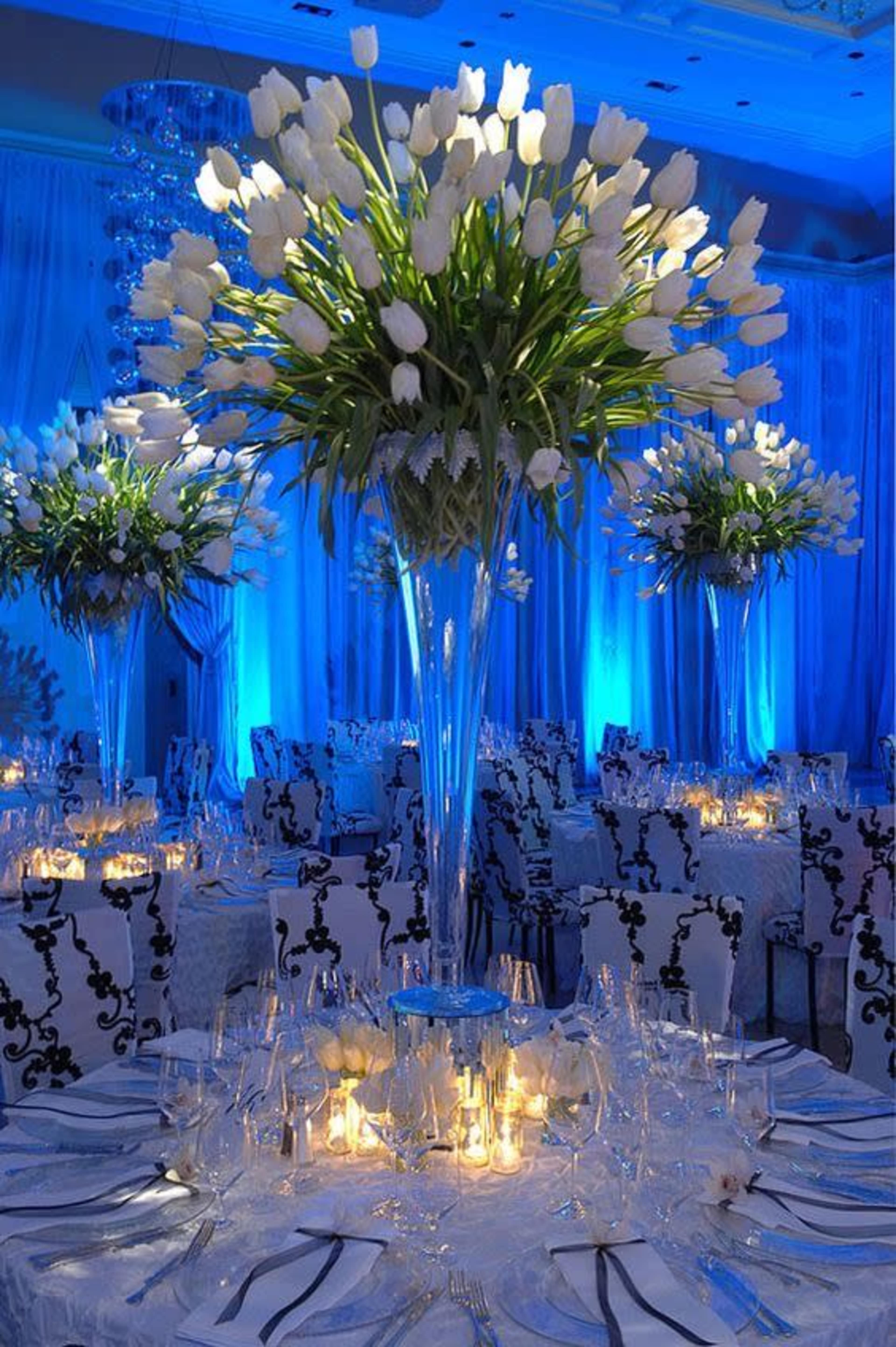 The image shows a decorated banquet hall with tall floral arrangements and illuminated tables set for an event.