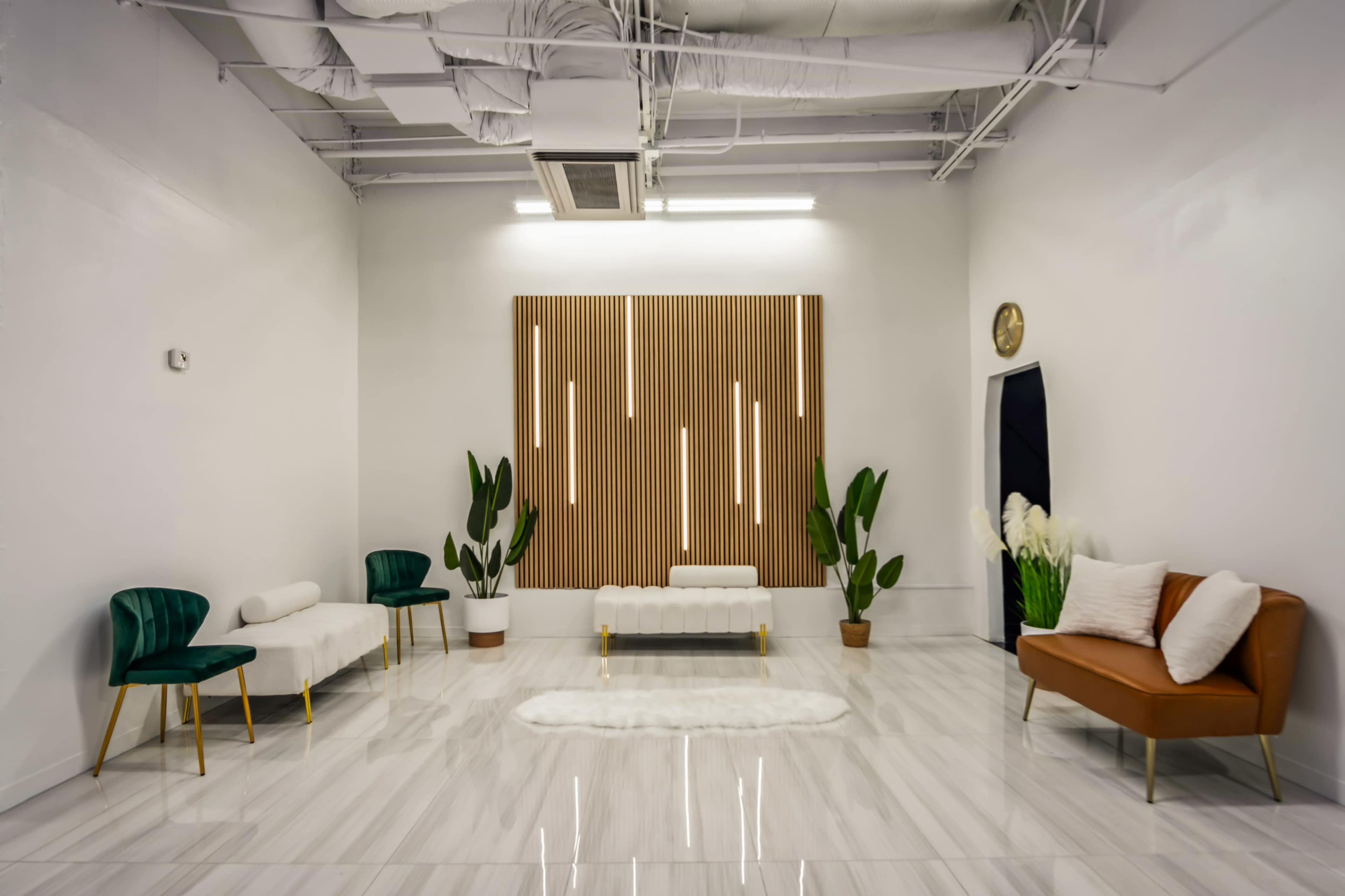 A modern, minimalist interior featuring white walls, a glossy floor, a wooden accent wall with vertical lights, and several pieces of furniture, including two green chairs, a brown sofa, and potted plants.