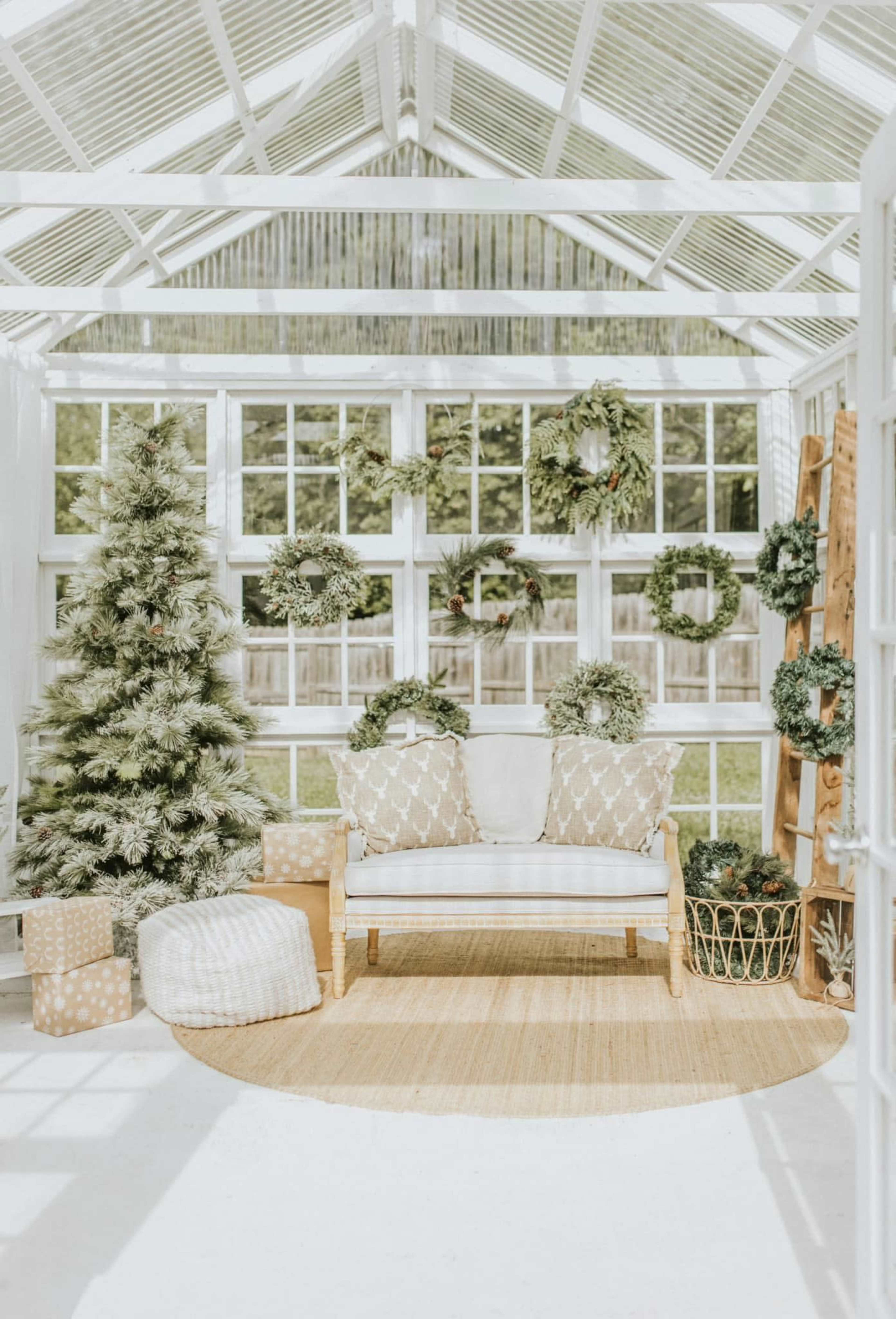 A bright conservatory features a white sofa, decorated with patterned cushions, surrounded by greenery, wreaths, and holiday gifts.