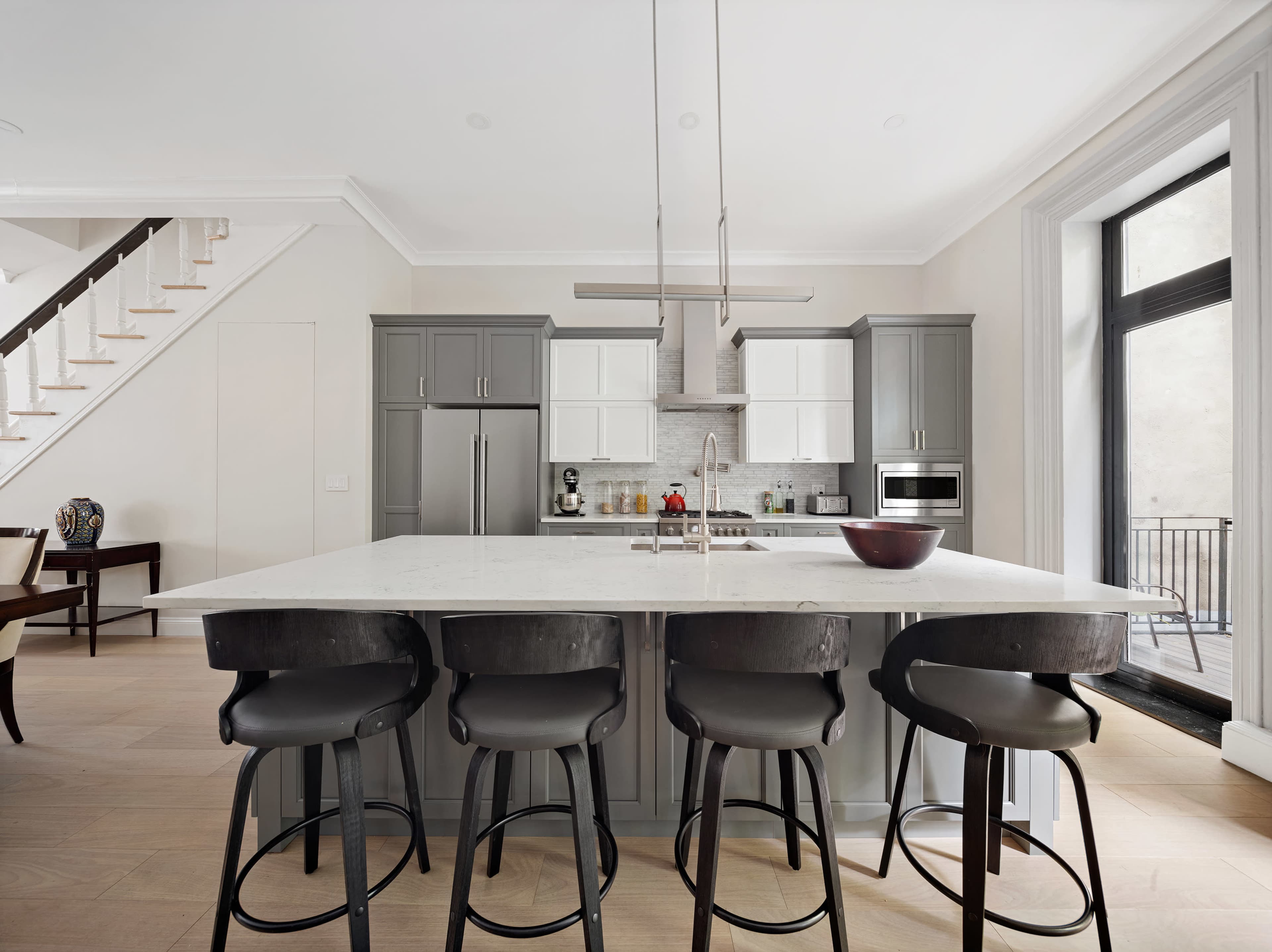 A modern kitchen features gray cabinetry, a large white marble island with black bar stools, and stainless steel appliances.