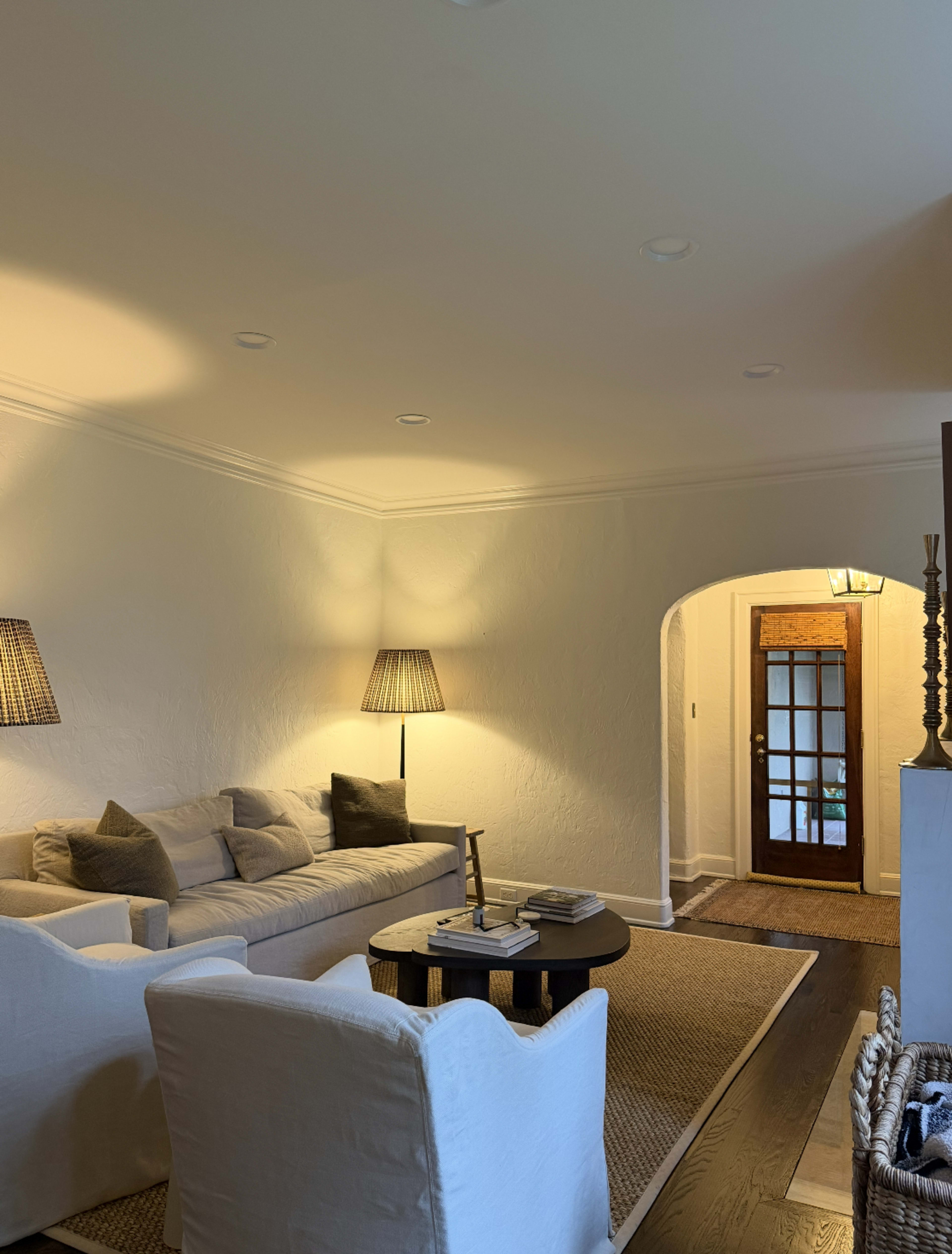 The image shows a cozy living room with a beige sofa, two white armchairs, a round coffee table, and a doorway leading to an entrance with a wooden door.
