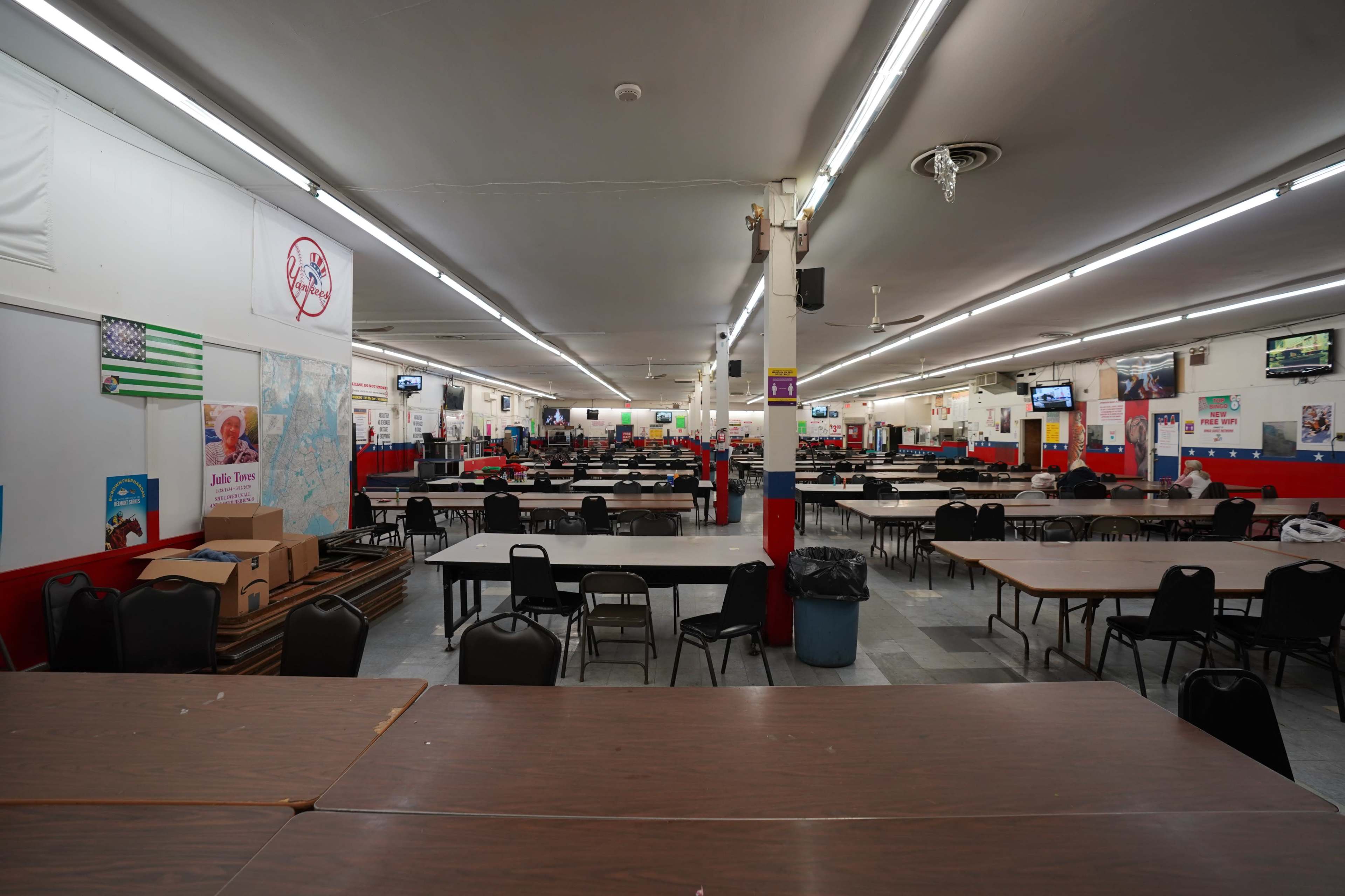The image shows an empty dining hall with rows of tables and chairs, illuminated by fluorescent lights.