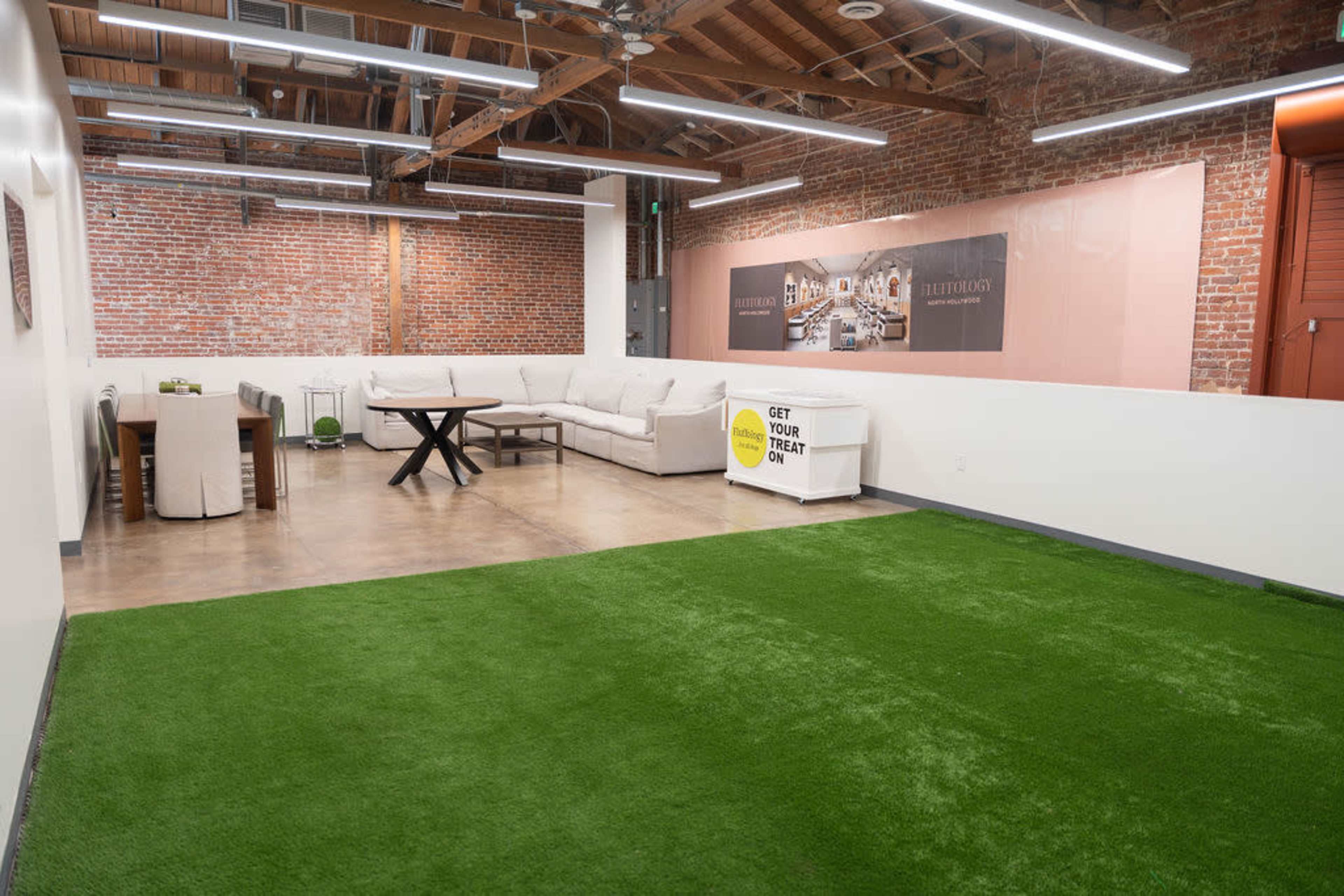 The image shows a spacious interior with exposed brick walls, a large sectional sofa, a dining table, and an area covered in green artificial turf.