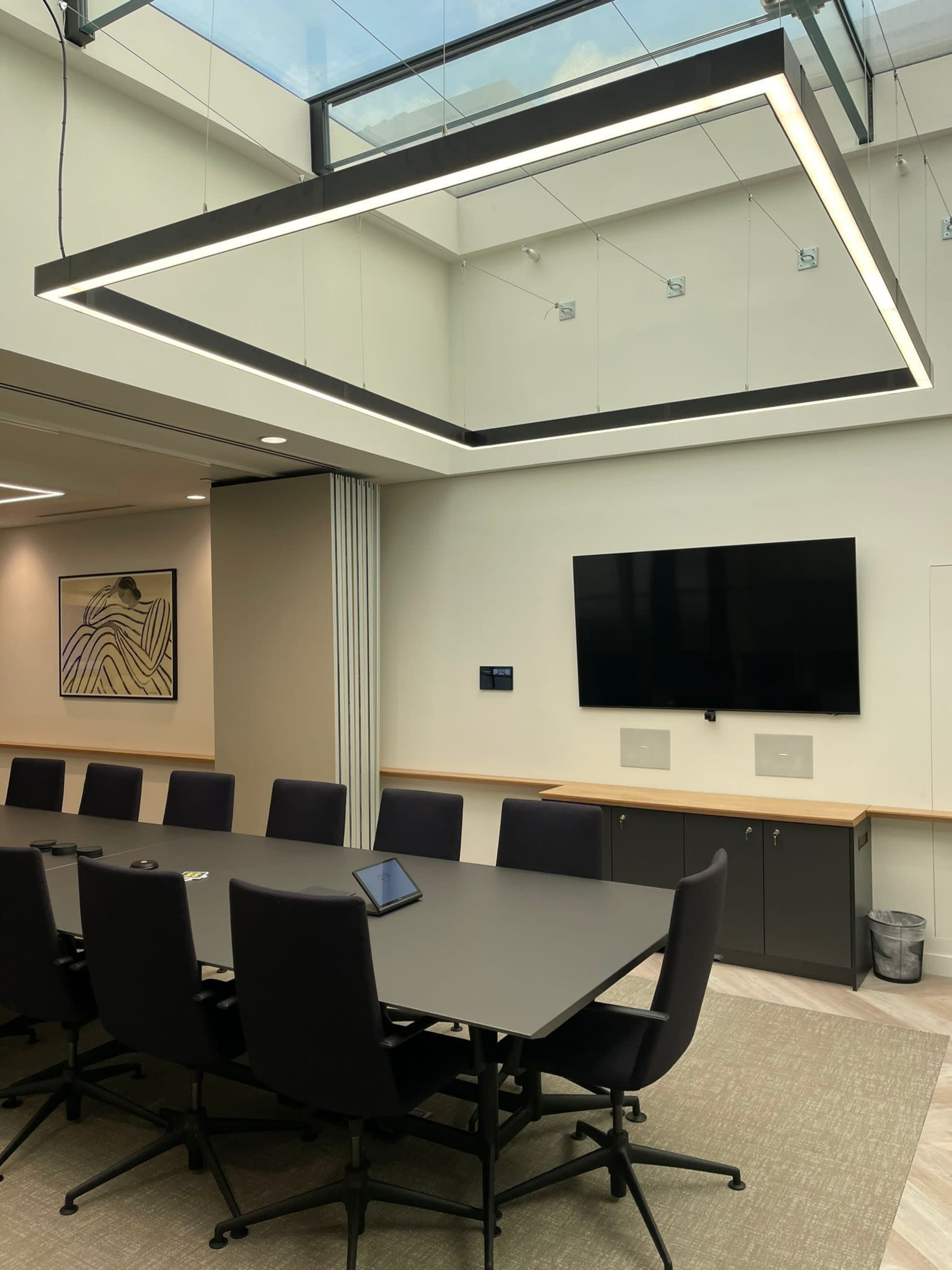A modern conference room features a large rectangular table surrounded by black chairs, with a ceiling-mounted light fixture and a screen on the wall.