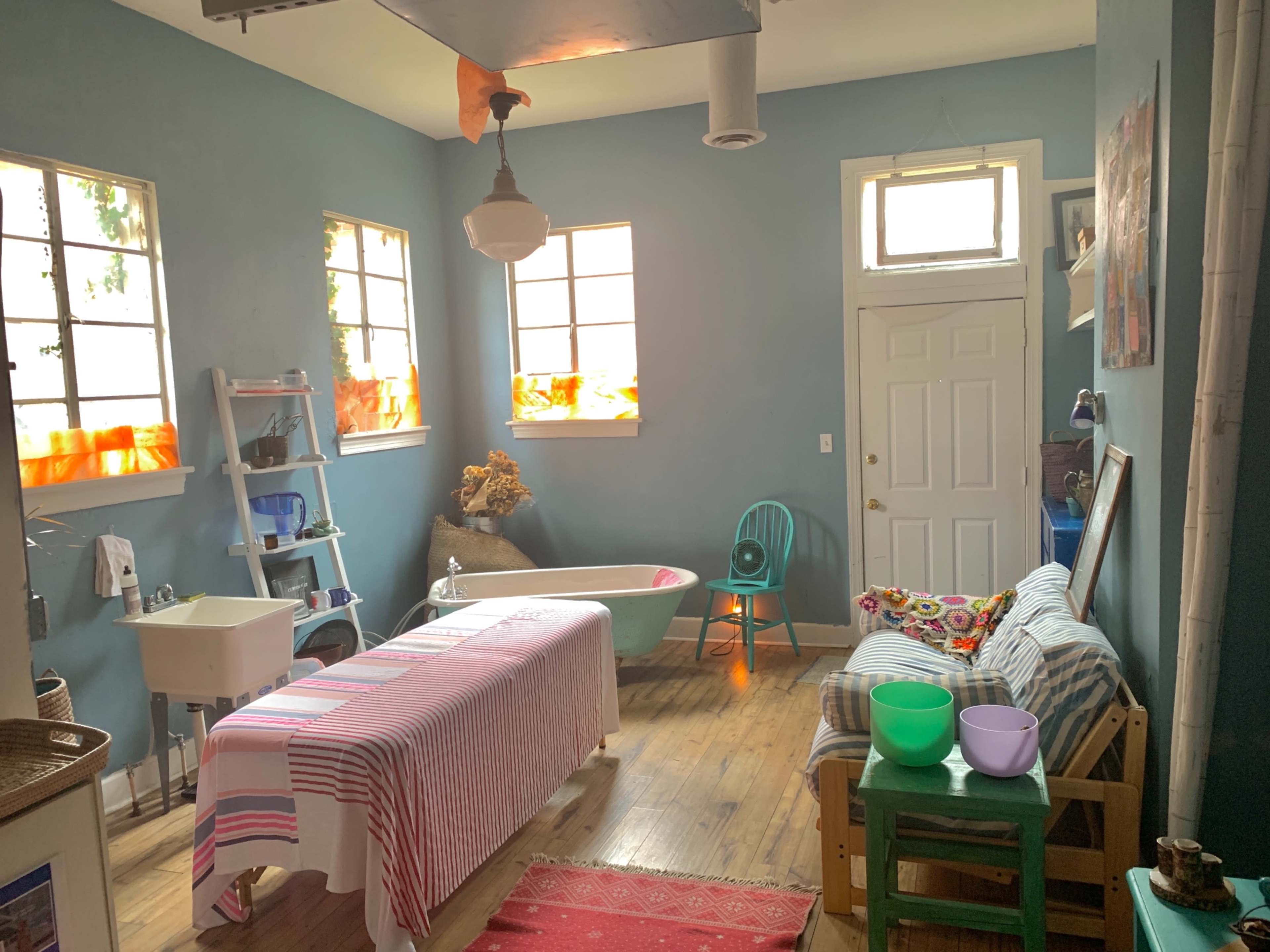 The room features a bathtub, a table covered with a striped cloth, a green chair, and various colorful objects against blue walls with windows letting in natural light.
