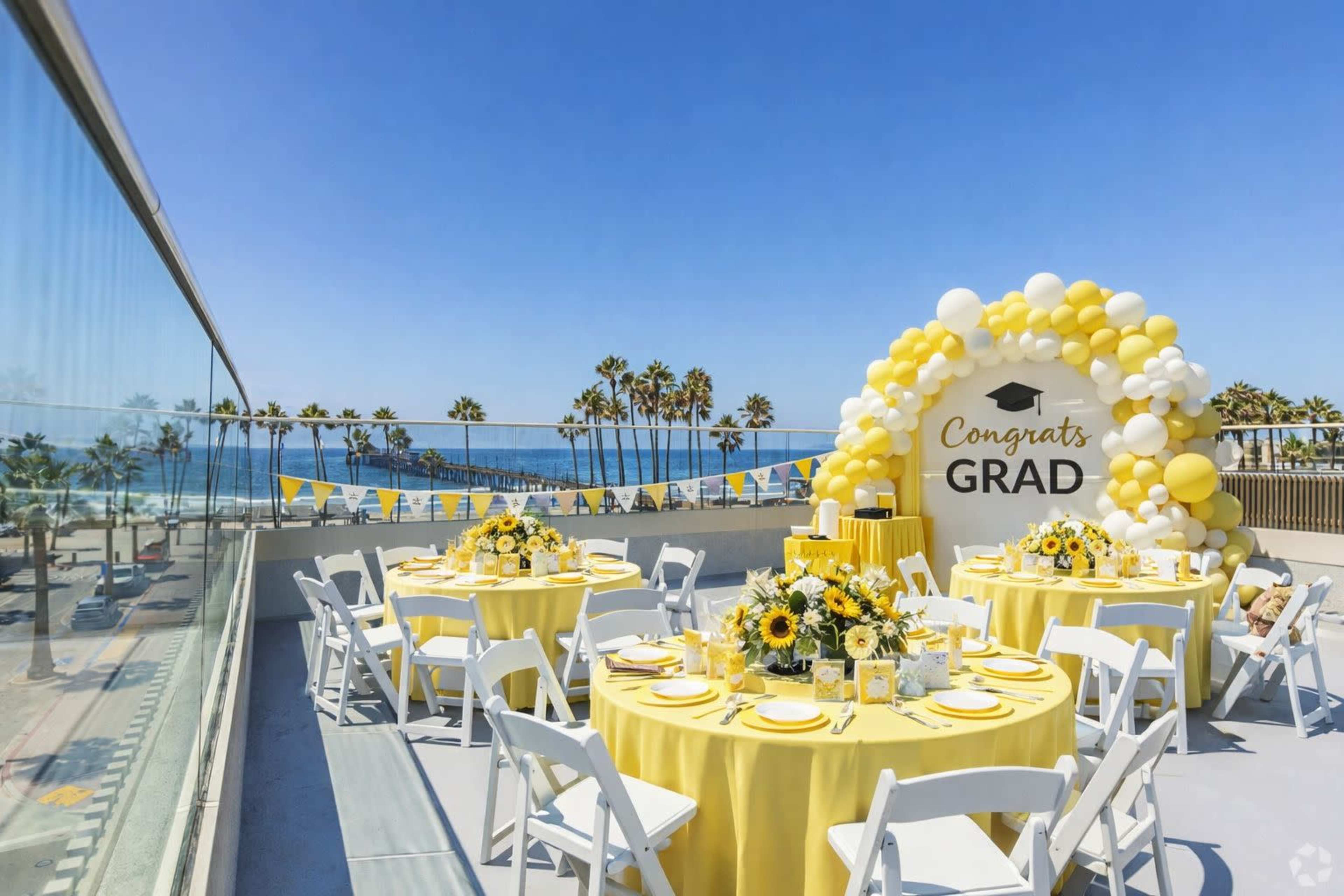 A rooftop celebration setup features round tables adorned with yellow tablecloths and sunflower centerpieces, with a large balloon display and a view of palm trees and the ocean in the background.