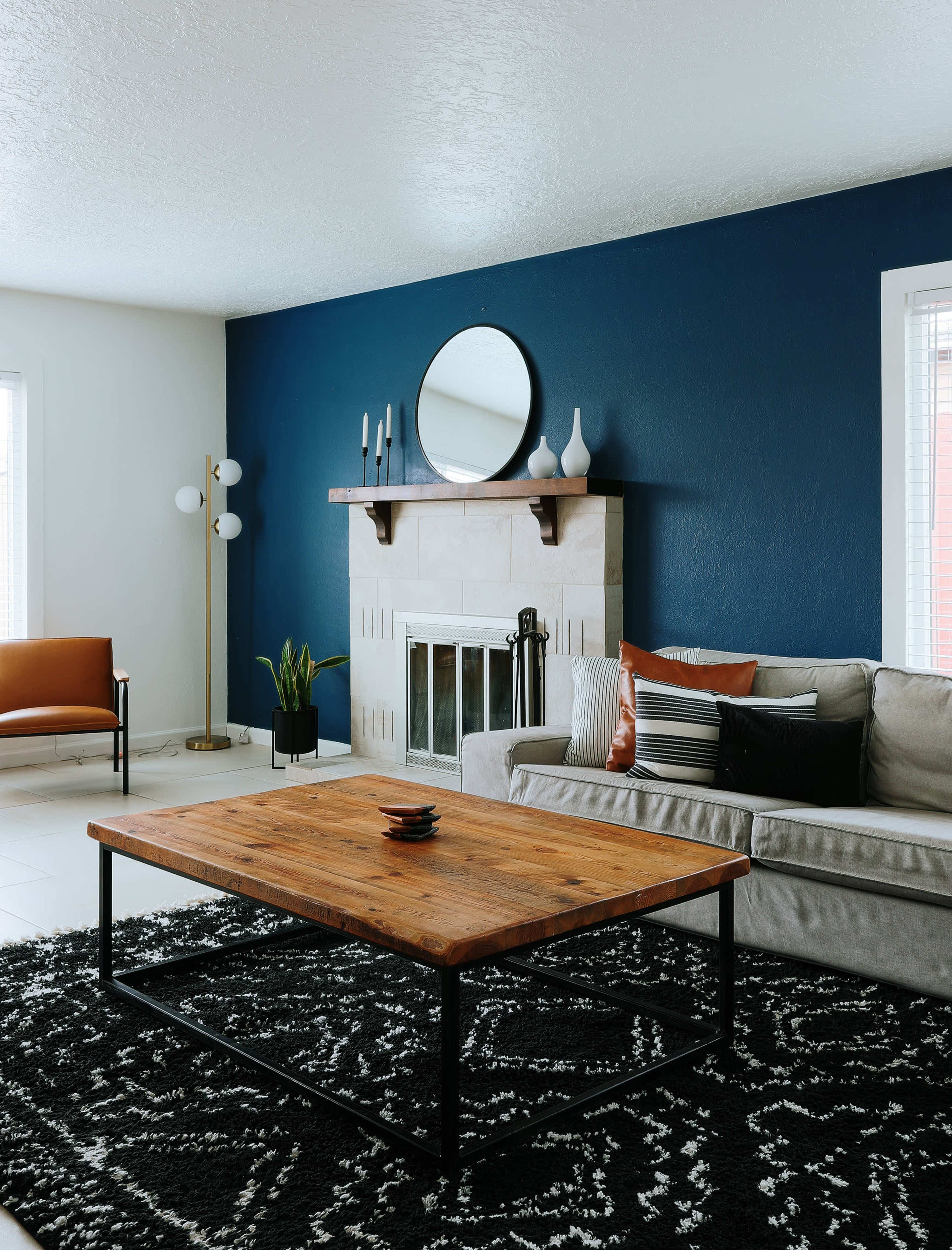 The living room features a wooden coffee table on a patterned rug, a gray sofa with pillows, a fireplace with a round mirror above it, and a chair beside a floor lamp, all accented by a dark blue wall.