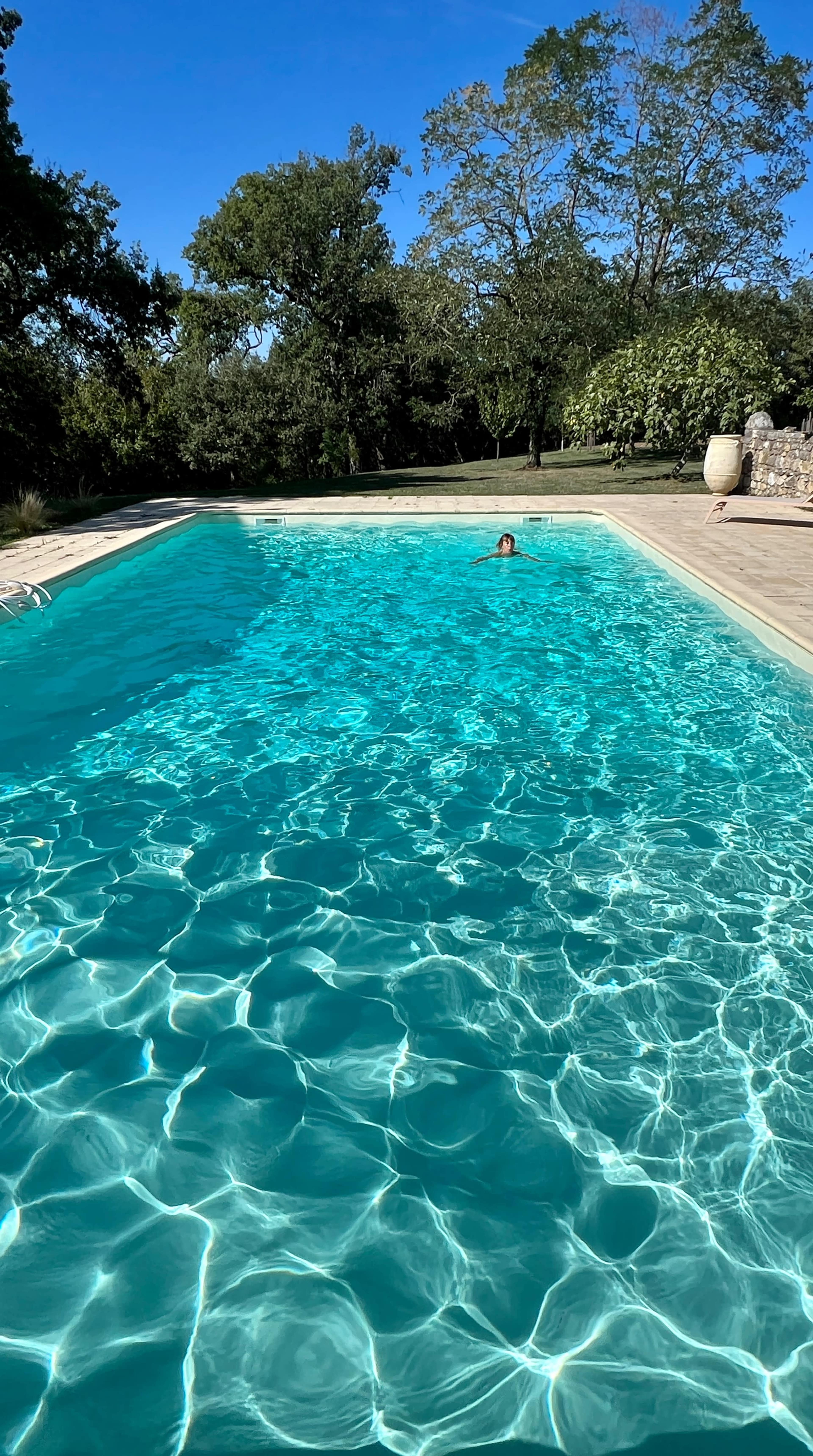 A person is swimming in a clear blue swimming pool surrounded by trees and a patio area.