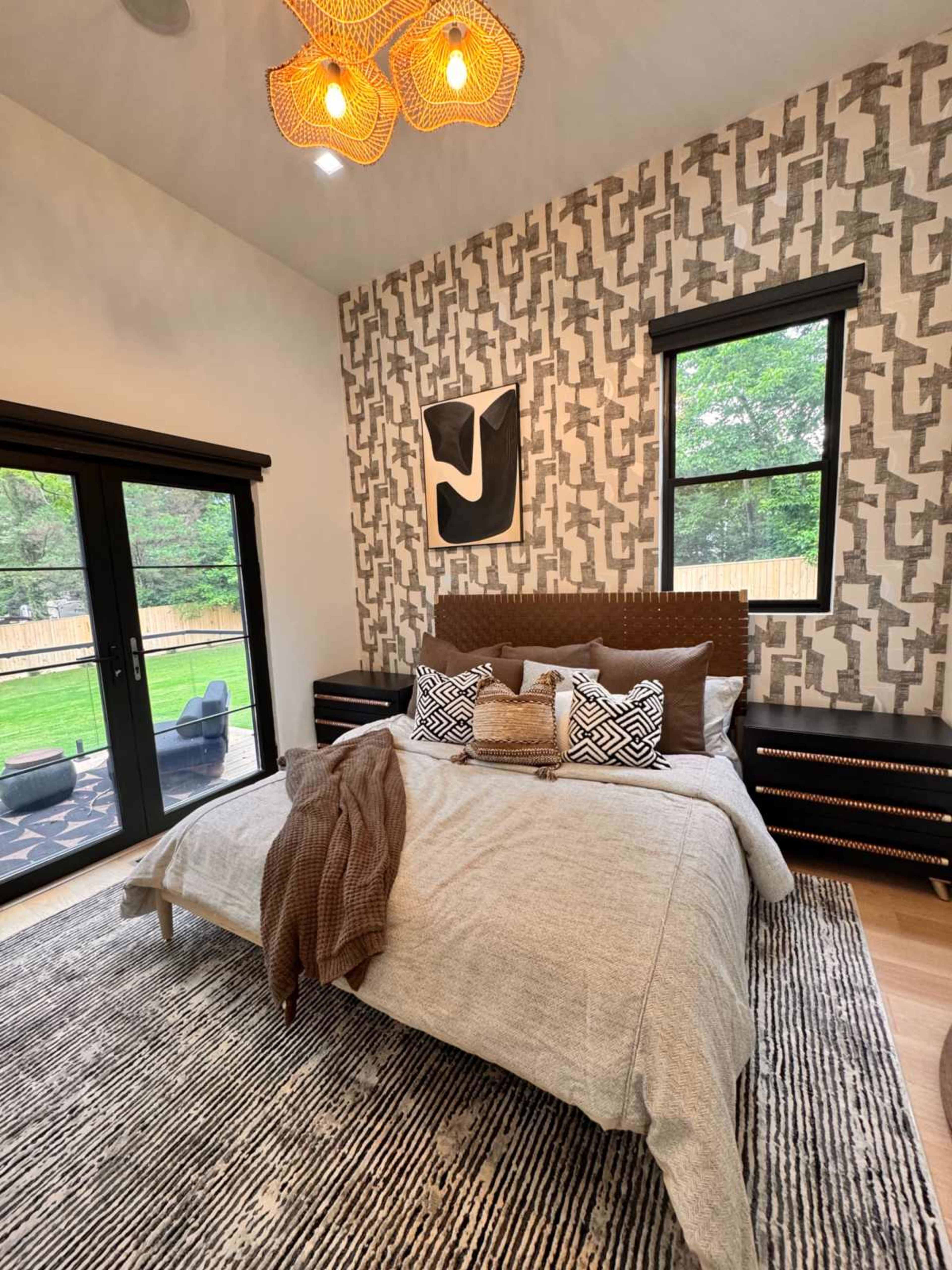 The image shows a modern bedroom with a large bed, patterned wallpaper, and stylish lighting fixtures, overlooking a grassy yard through glass doors.
