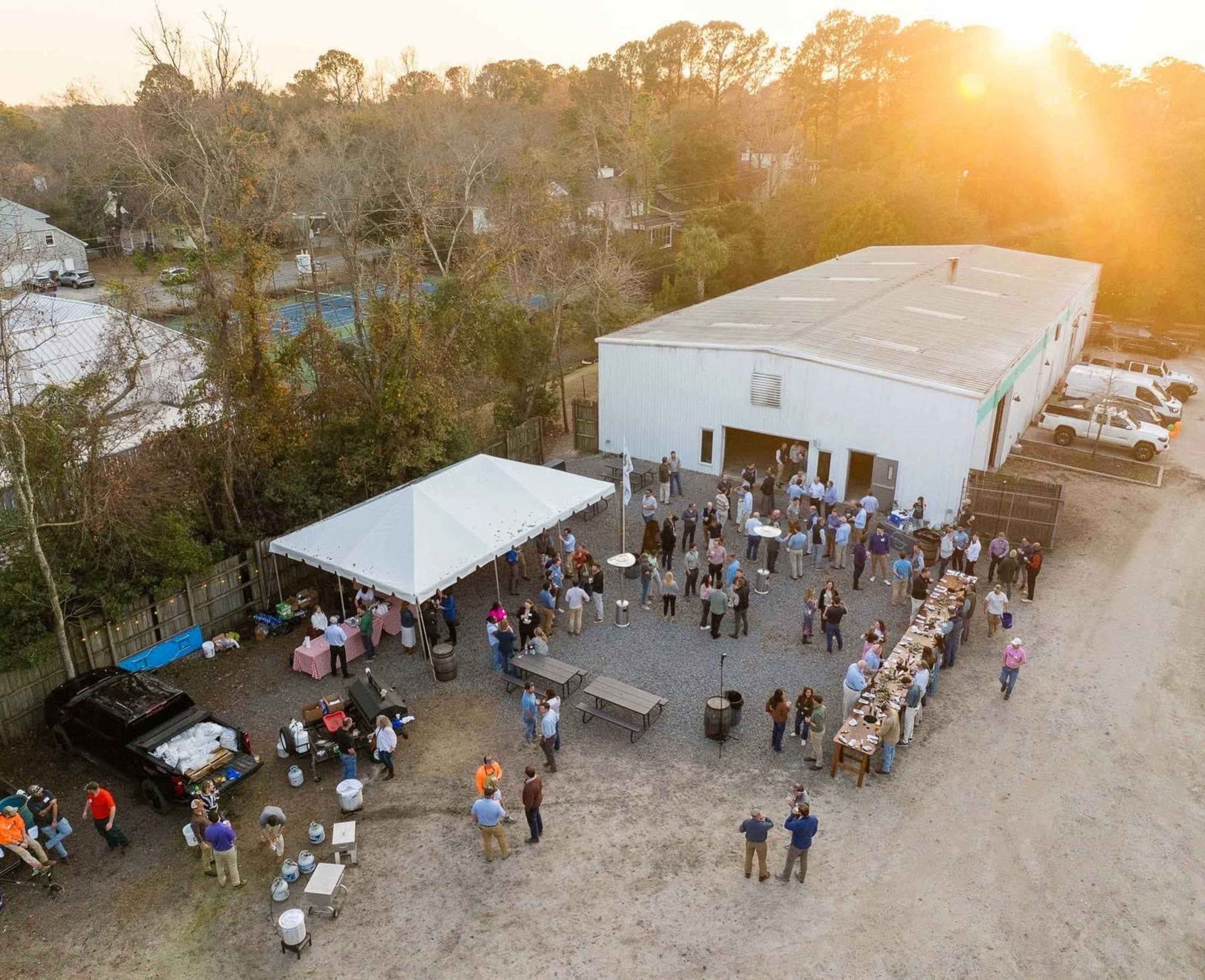A large gathering of people socializes under a tent and around tables outside a white industrial building during sunset.