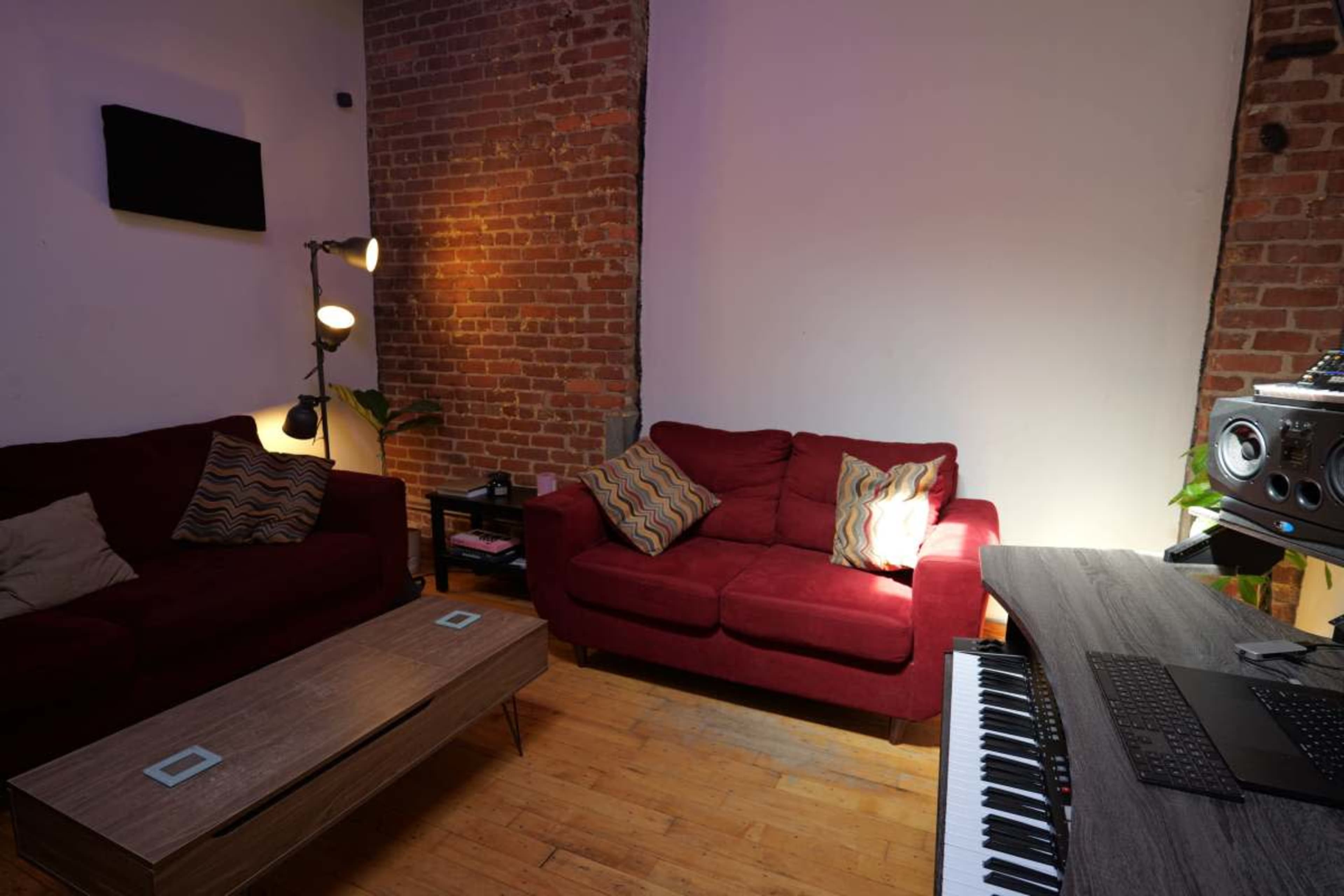 The image shows a cozy room featuring two red sofas, a wooden coffee table, and a music production setup with a keyboard and speakers.