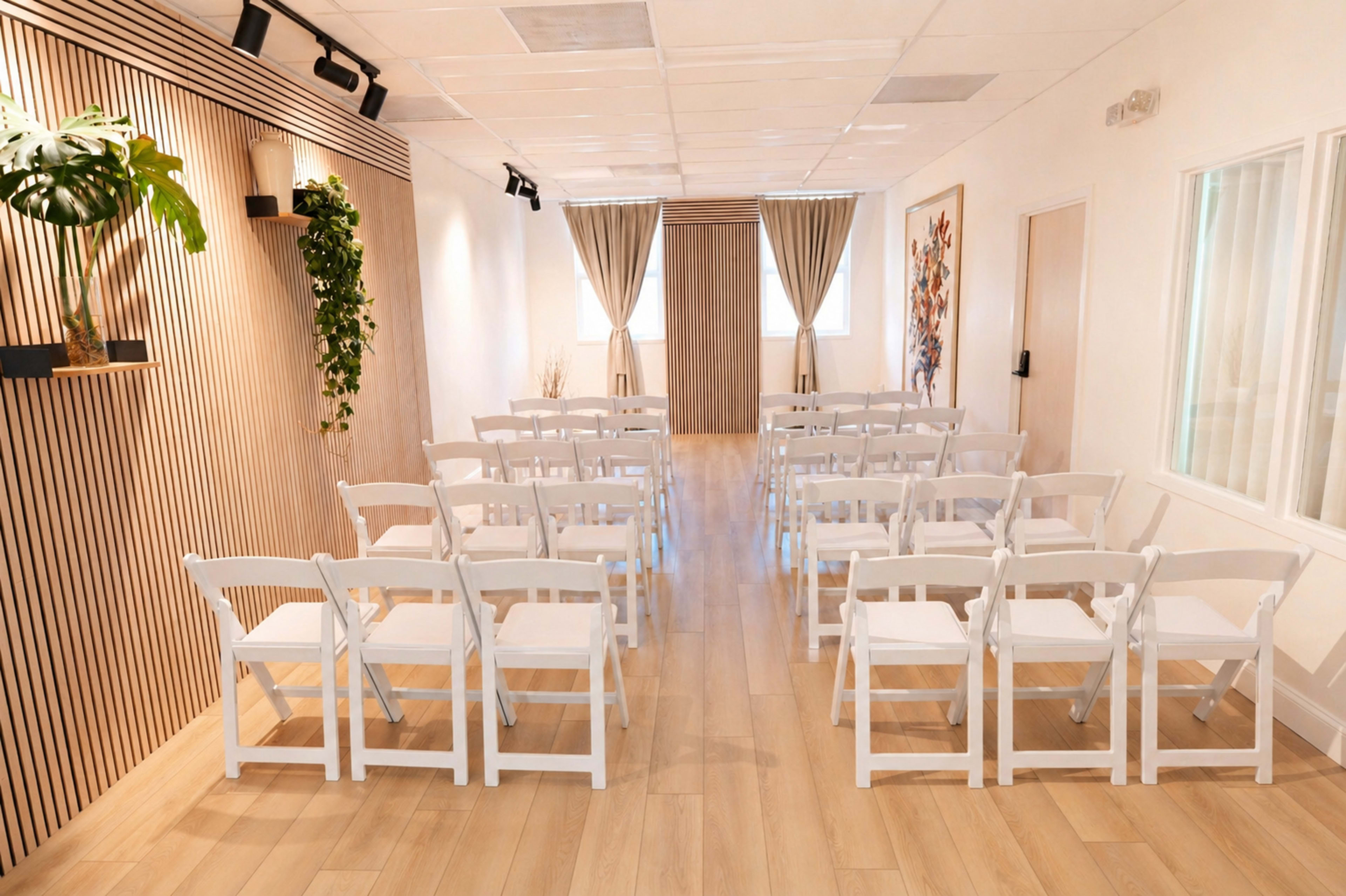 The image shows a bright interior space set up with rows of white folding chairs facing a wooden door, complemented by plants and decorative wall elements.