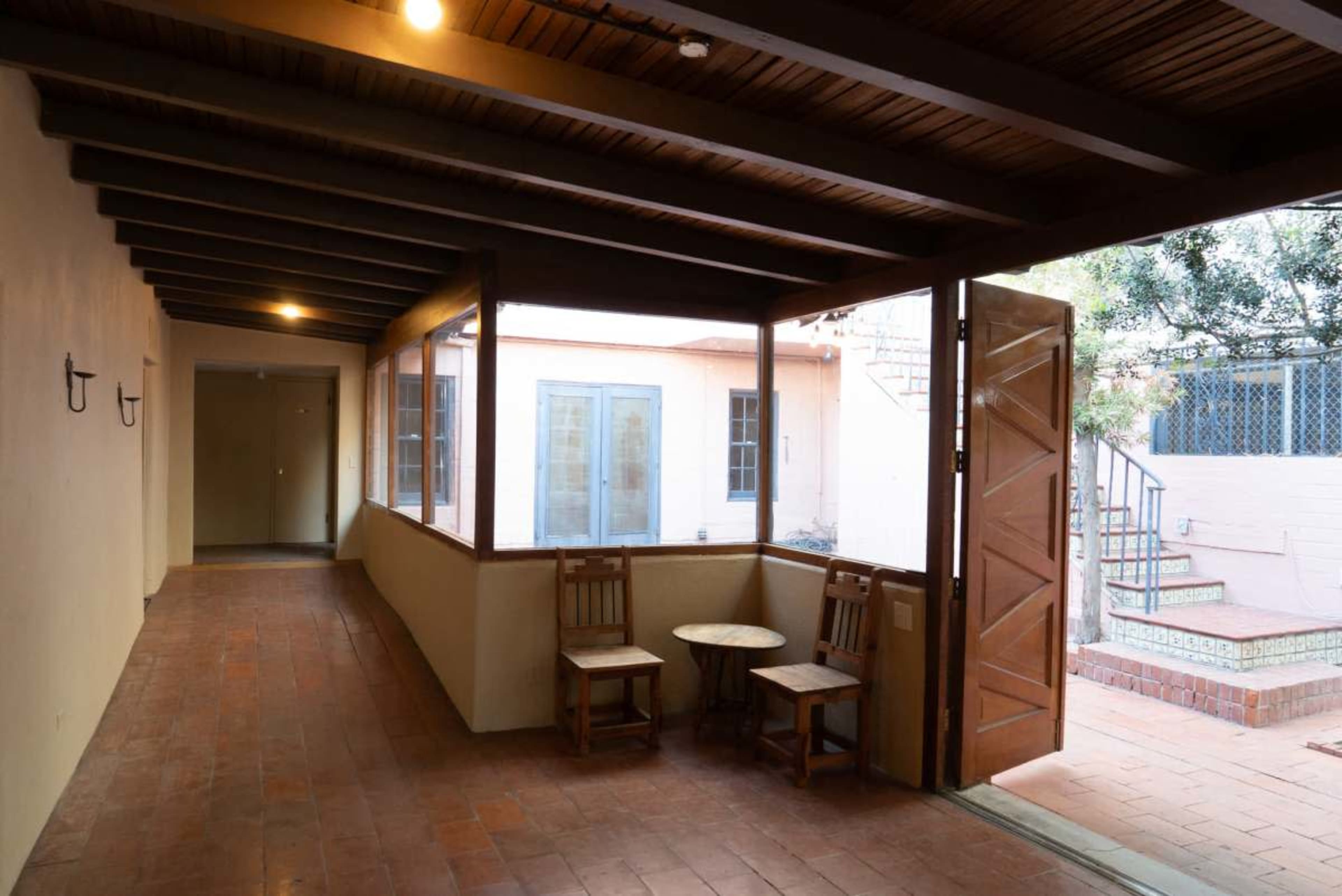 The image shows a covered hallway with wooden beams, leading to a door on one side and a view of an outdoor area with steps on the other.