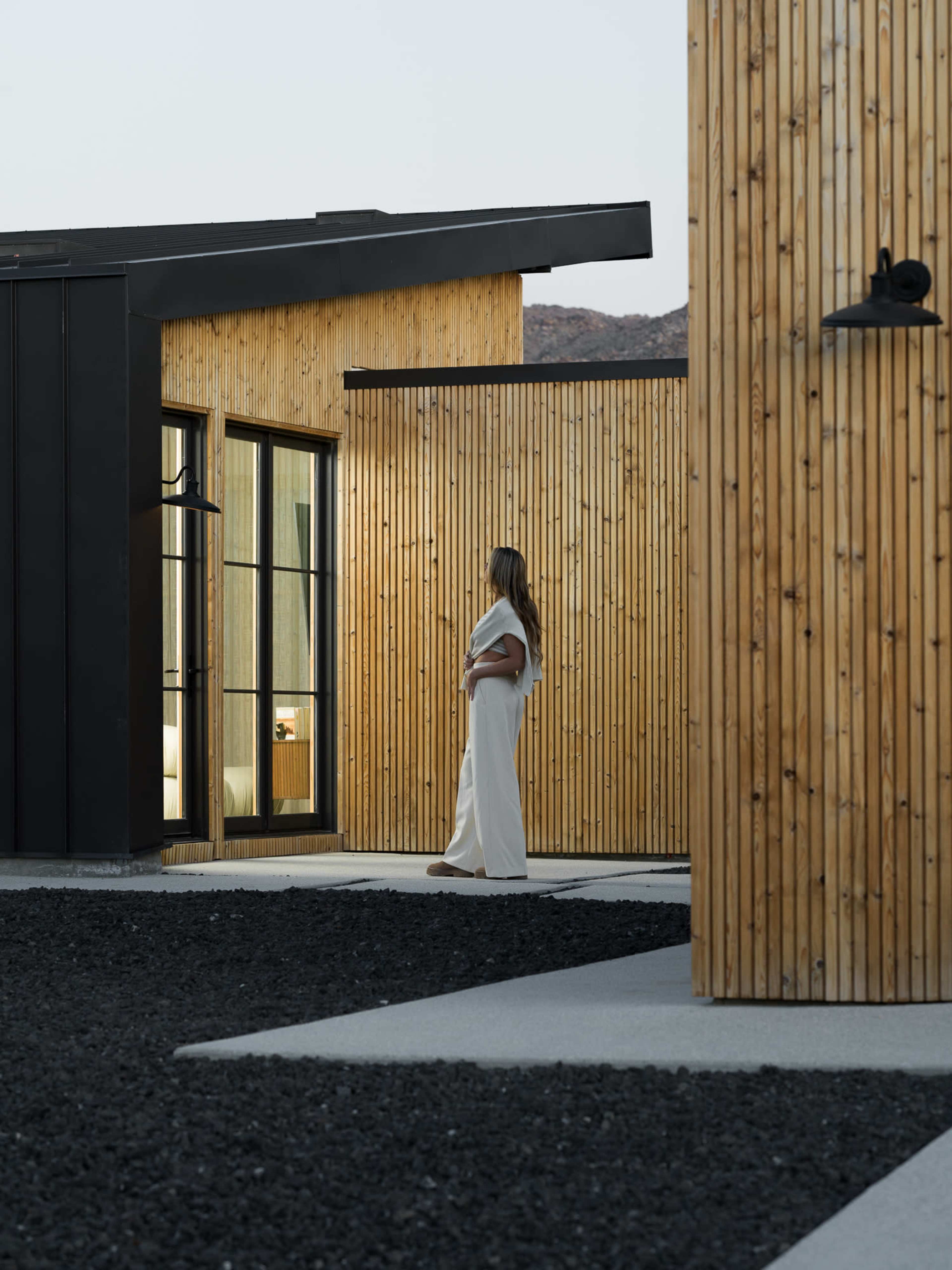 A woman in a white dress stands next to a modern wooden facade of a building, surrounded by a gravel landscape.