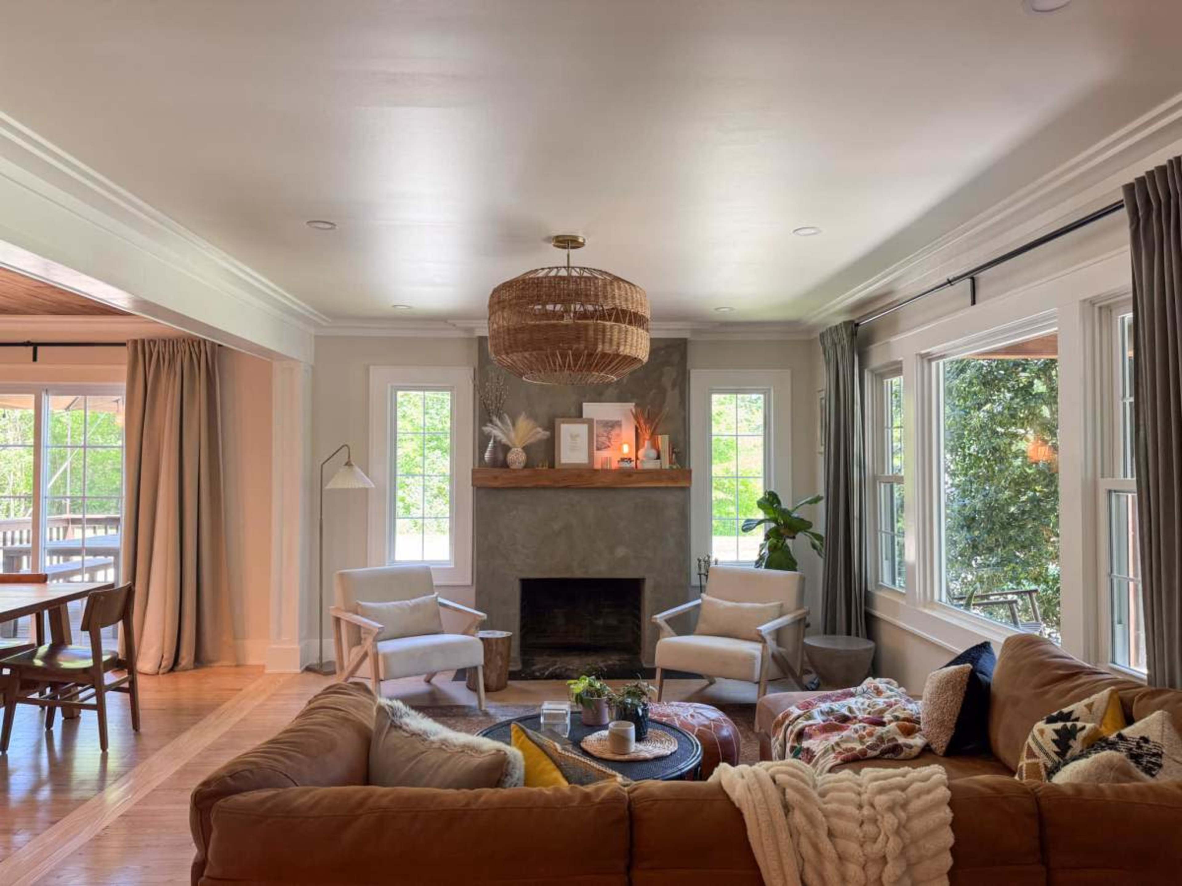 The image shows a cozy living room with a sofa, two armchairs, a stone fireplace, and large windows allowing natural light to enter.