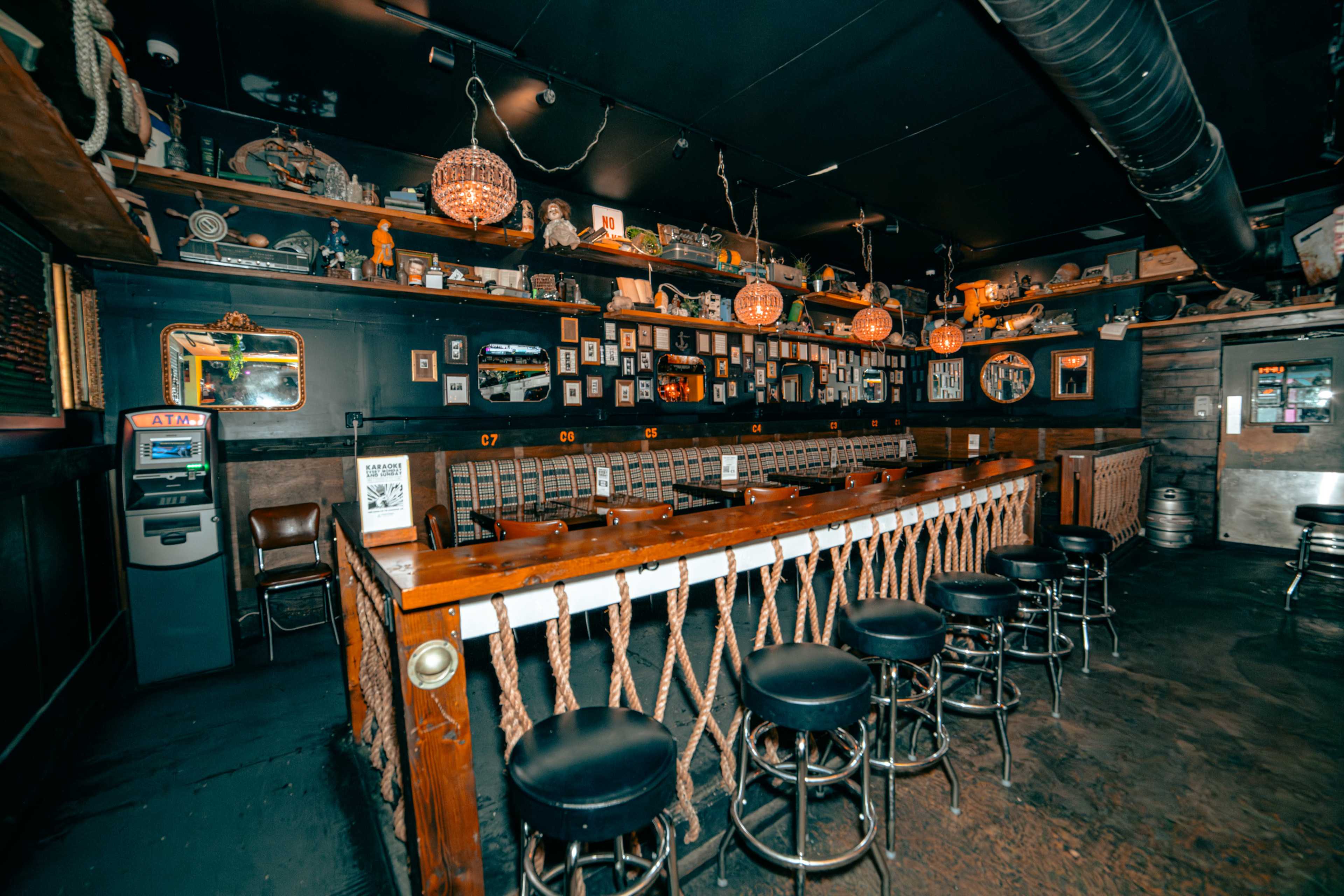 The image shows a dimly lit bar with a long wooden counter, bar stools, and shelves decorated with various items and memorabilia.