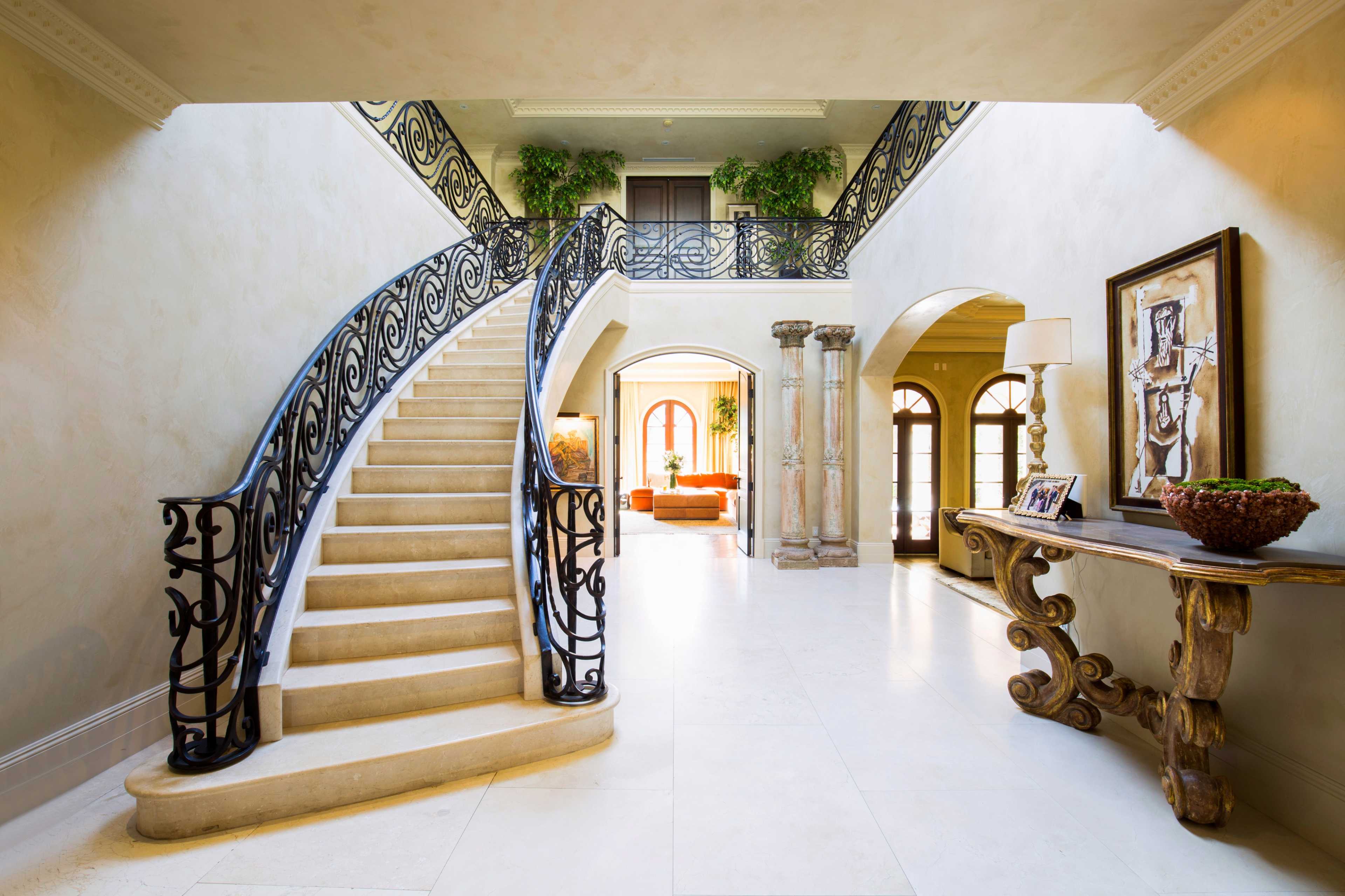 A spacious foyer with a sweeping staircase, elegant iron railing, and natural light illuminating the room filled with plants and decorative furnishings.