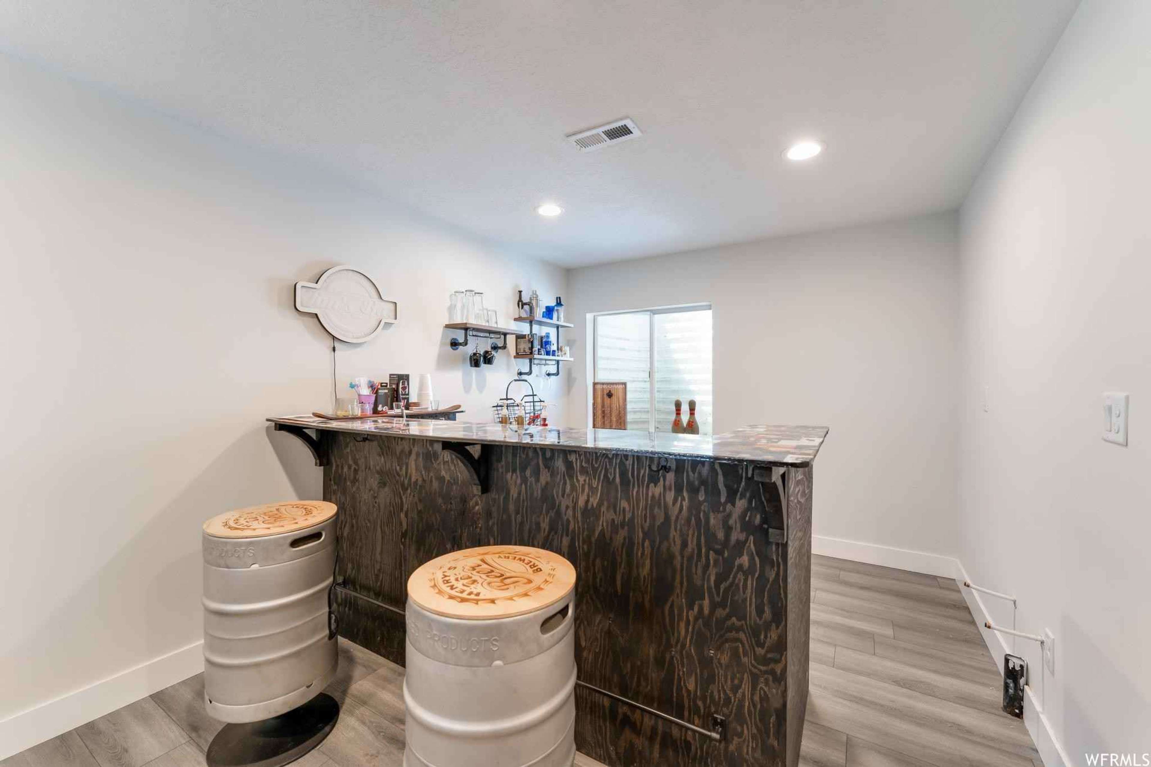 A modern home bar area with a dark wood counter, two barrel-style stools, and shelves holding various bottles and glassware.