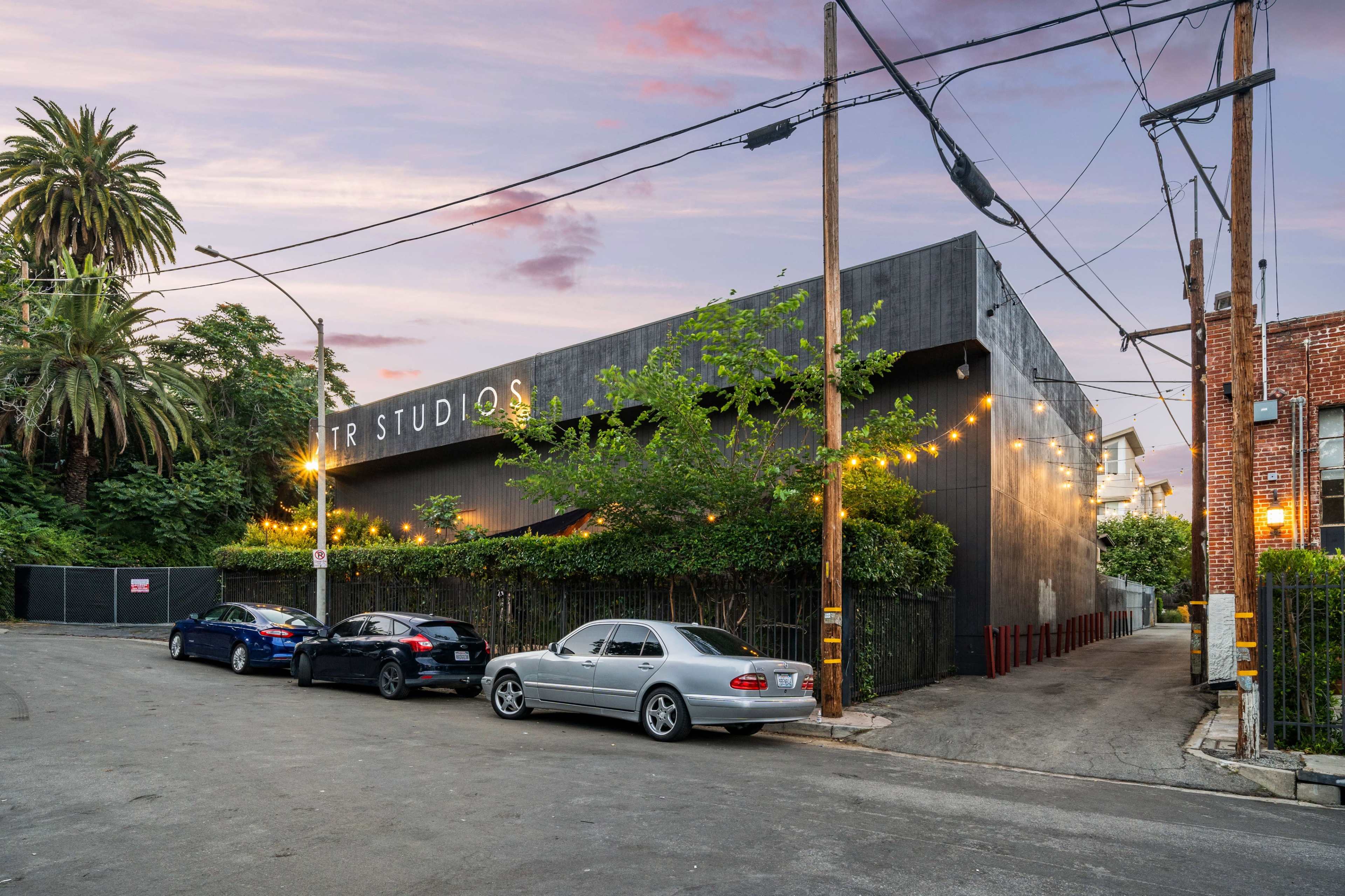 A modern building named "TR Studios" is situated at the corner of a street, surrounded by palm trees and string lights.