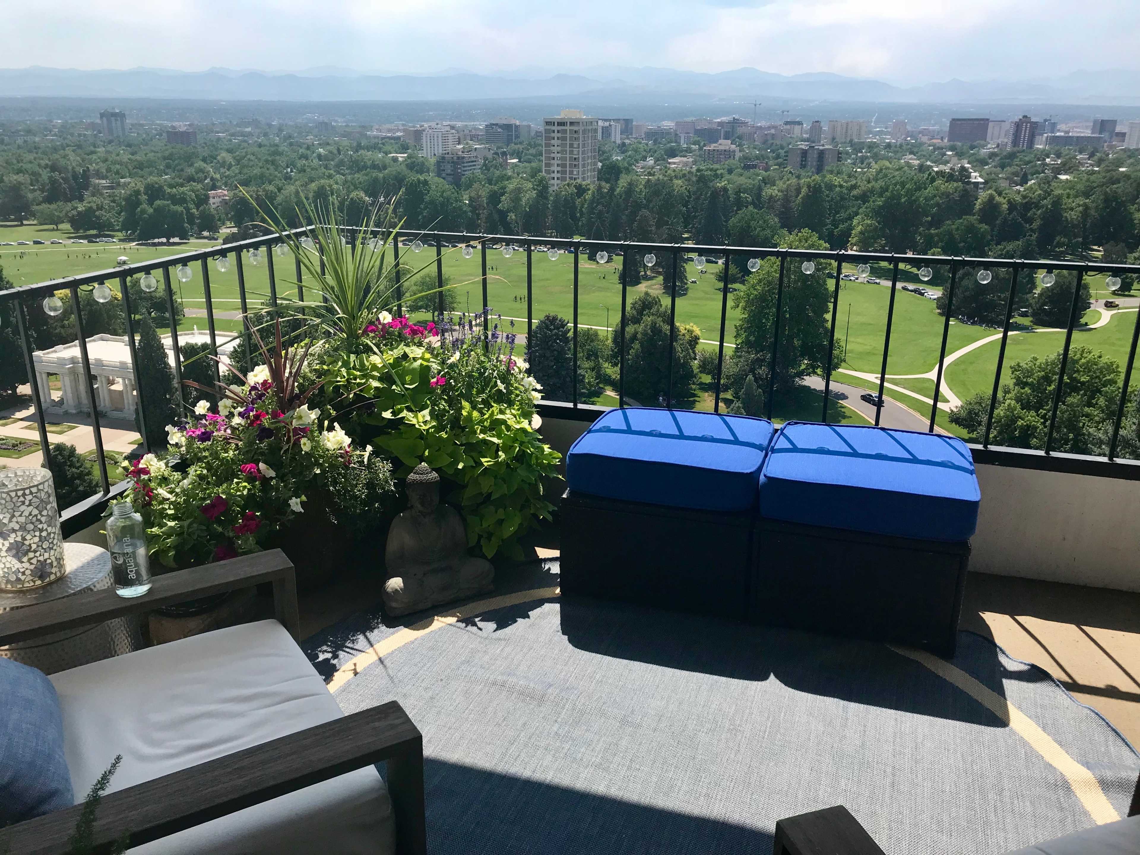 A balcony patio features blue cushioned seating, potted plants, and a scenic view of a city and mountains in the background.