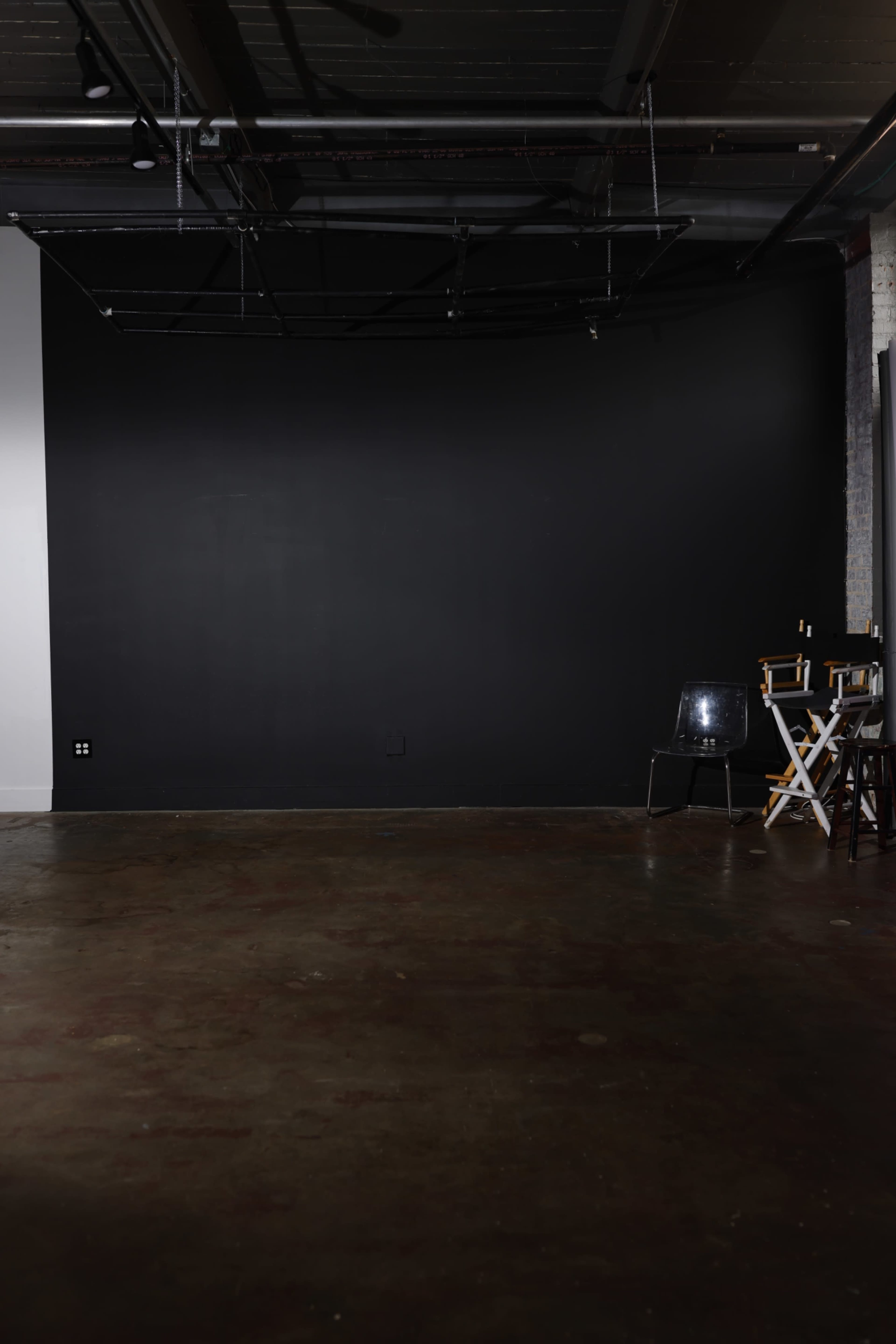 The image shows an empty studio space featuring a black wall, a polished concrete floor, and a director's chair next to a small table.