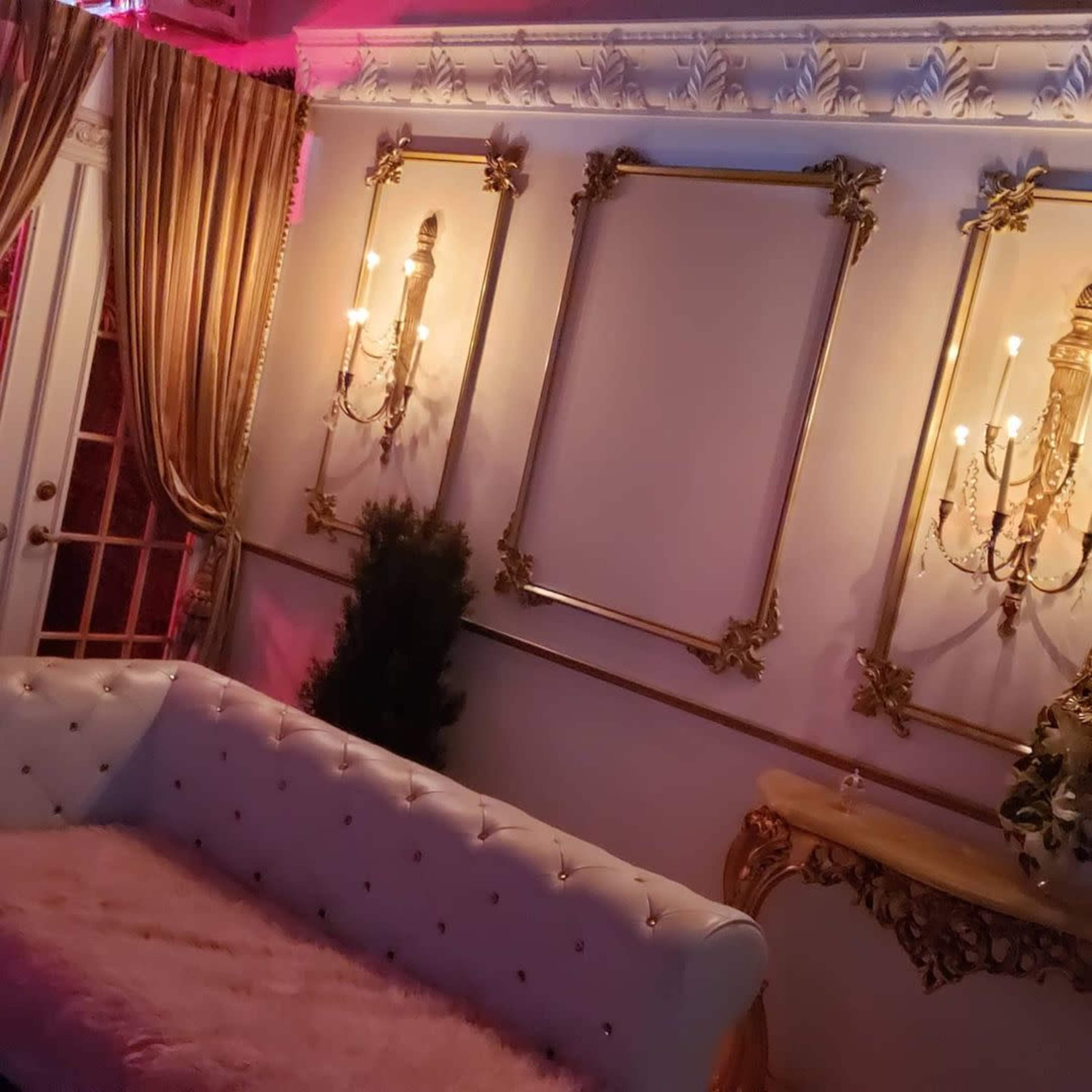An elegantly decorated interior featuring a tufted white sofa, ornate wall sconces, and gold-framed wall panels with curtains in the background.