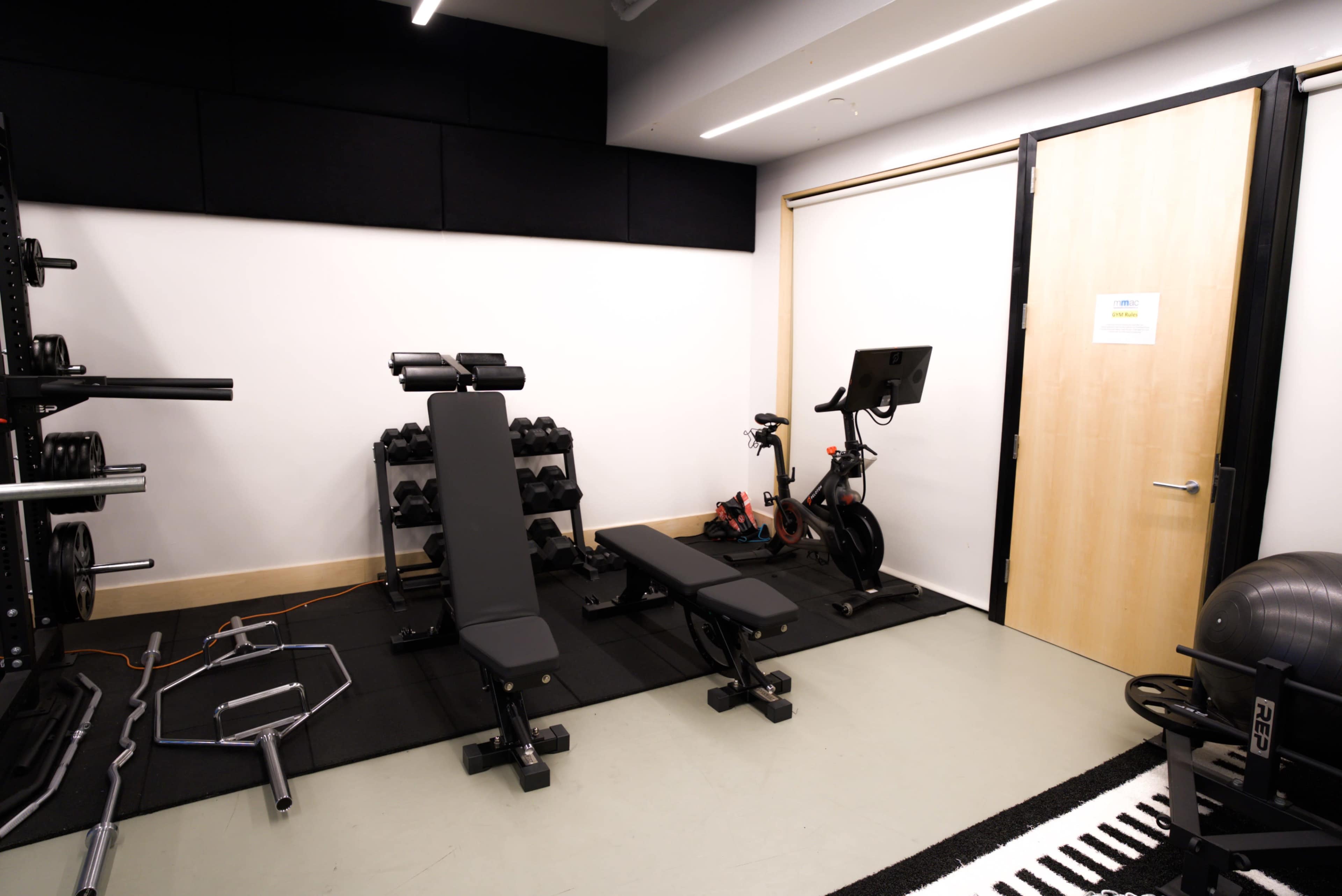 A compact fitness room featuring weightlifting equipment, a bench, dumbbells, and a stationary bike near a door.