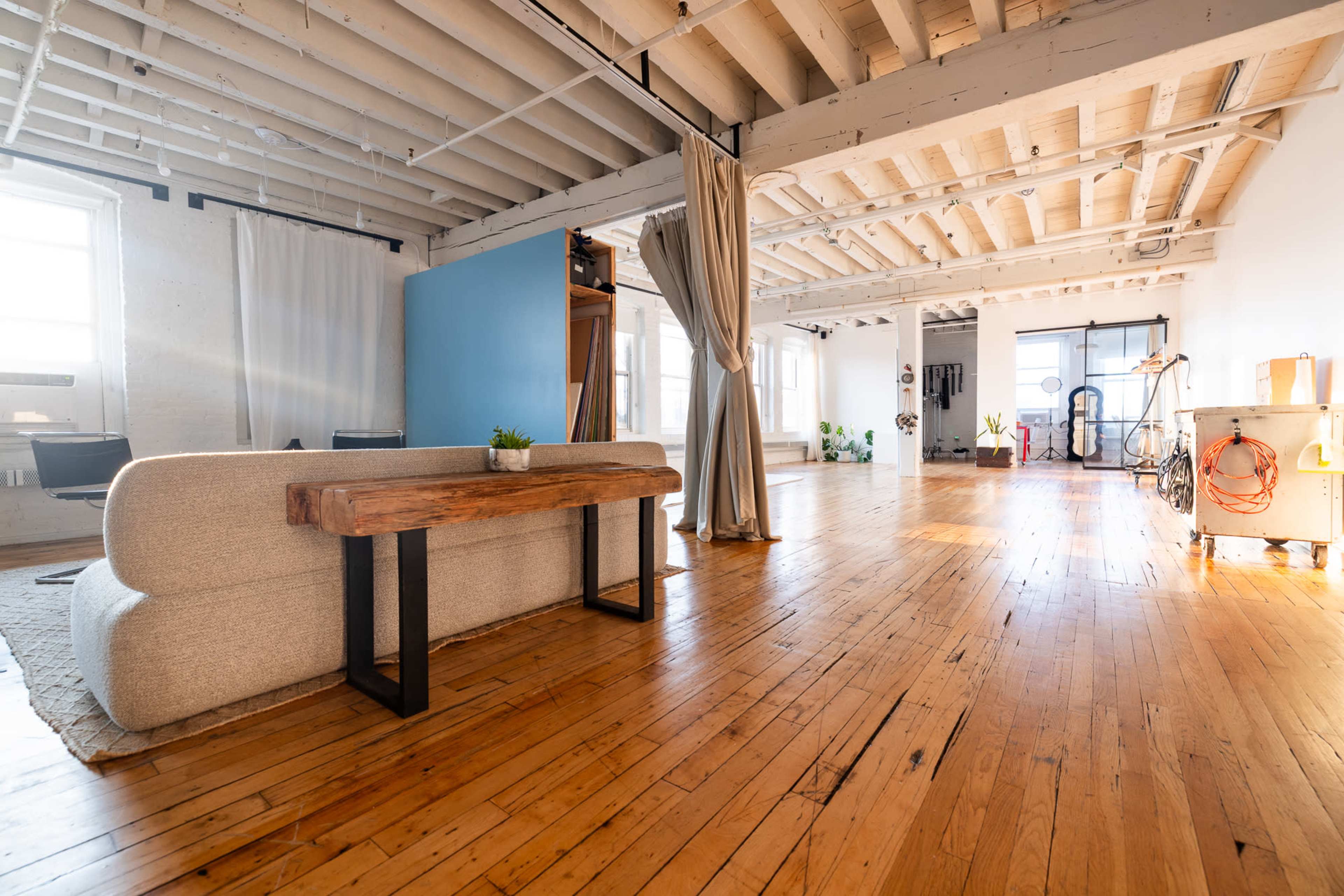 The image shows a spacious, well-lit loft with wooden floors, large windows, and minimalist furnishings, including a couch and a console table.